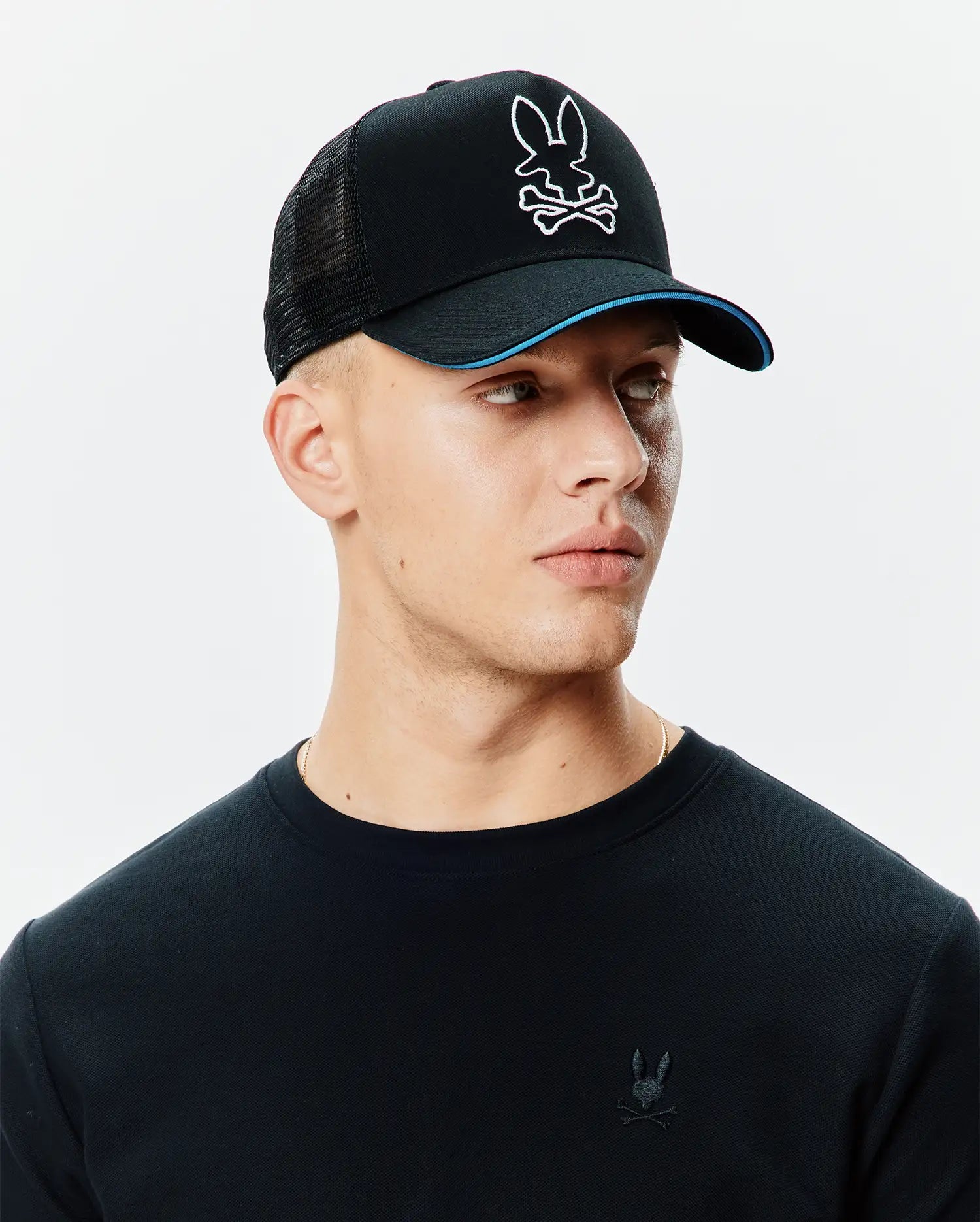 Black Mens Apollo Trucker Cap with mesh back, white embroidered Psycho Bunny skull logo, blue brim accent, and a black pique long-sleeve shirt with a small black embroidered Psycho Bunny logo on the left chest.