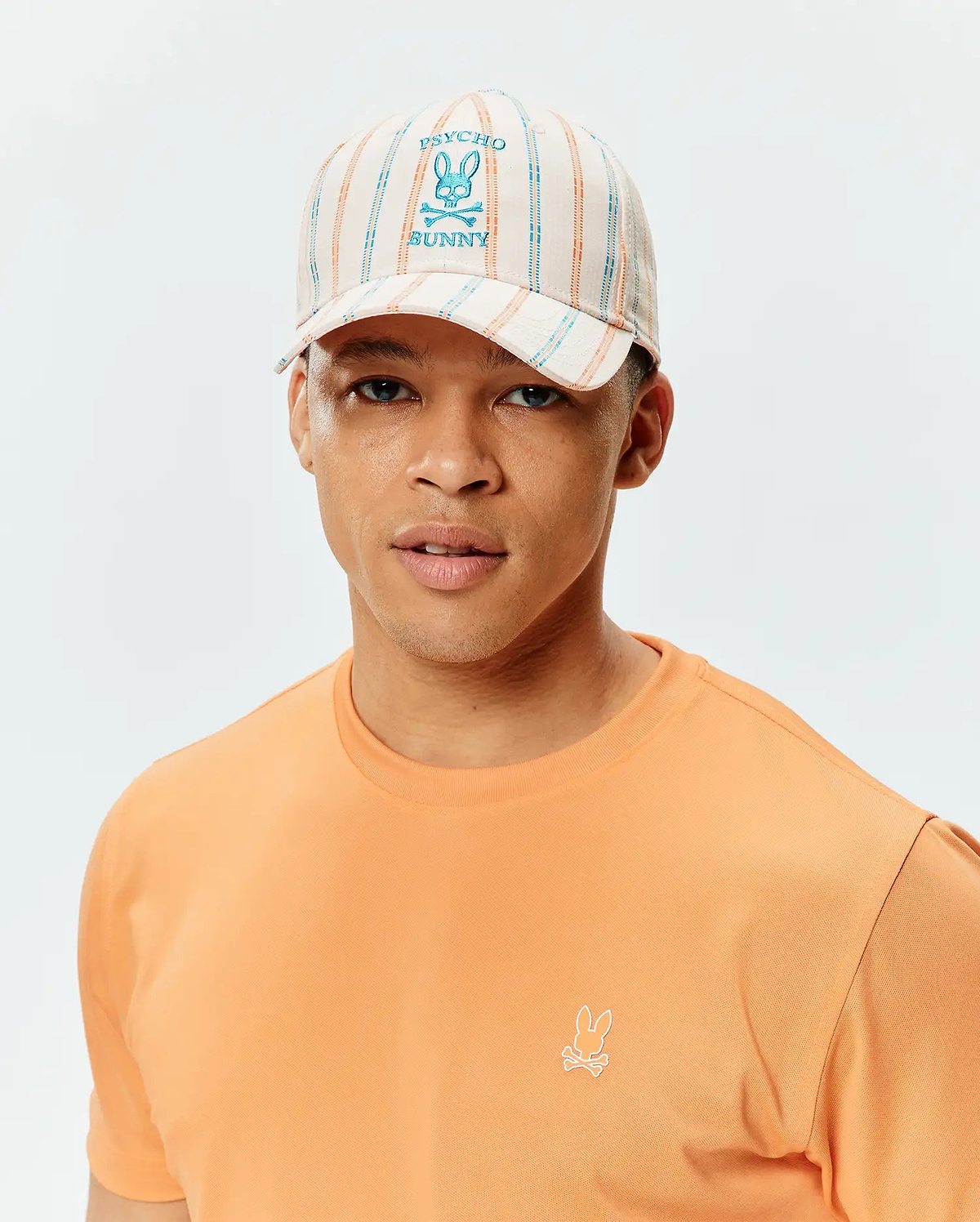 Mens Antoine Seersucker Baseball Cap with cream, blue, and orange stripes, featuring a large embroidered Psycho Bunny logo, paired with a peach classic fit jersey t-shirt with a small embroidered Psycho Bunny logo.