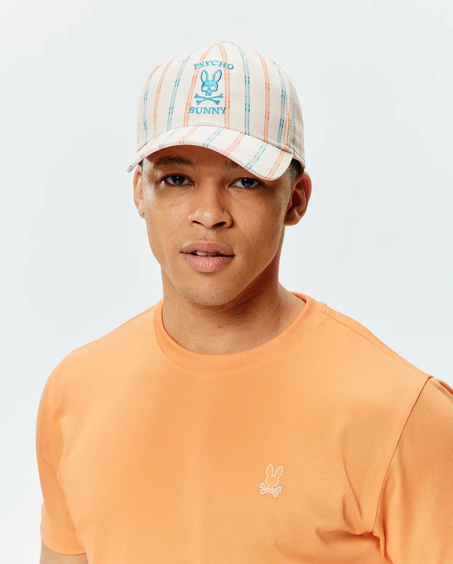 Mens Antoine Seersucker Baseball Cap with cream, blue, and orange stripes, featuring a large embroidered Psycho Bunny logo, paired with a peach classic fit jersey t-shirt with a small embroidered Psycho Bunny logo.