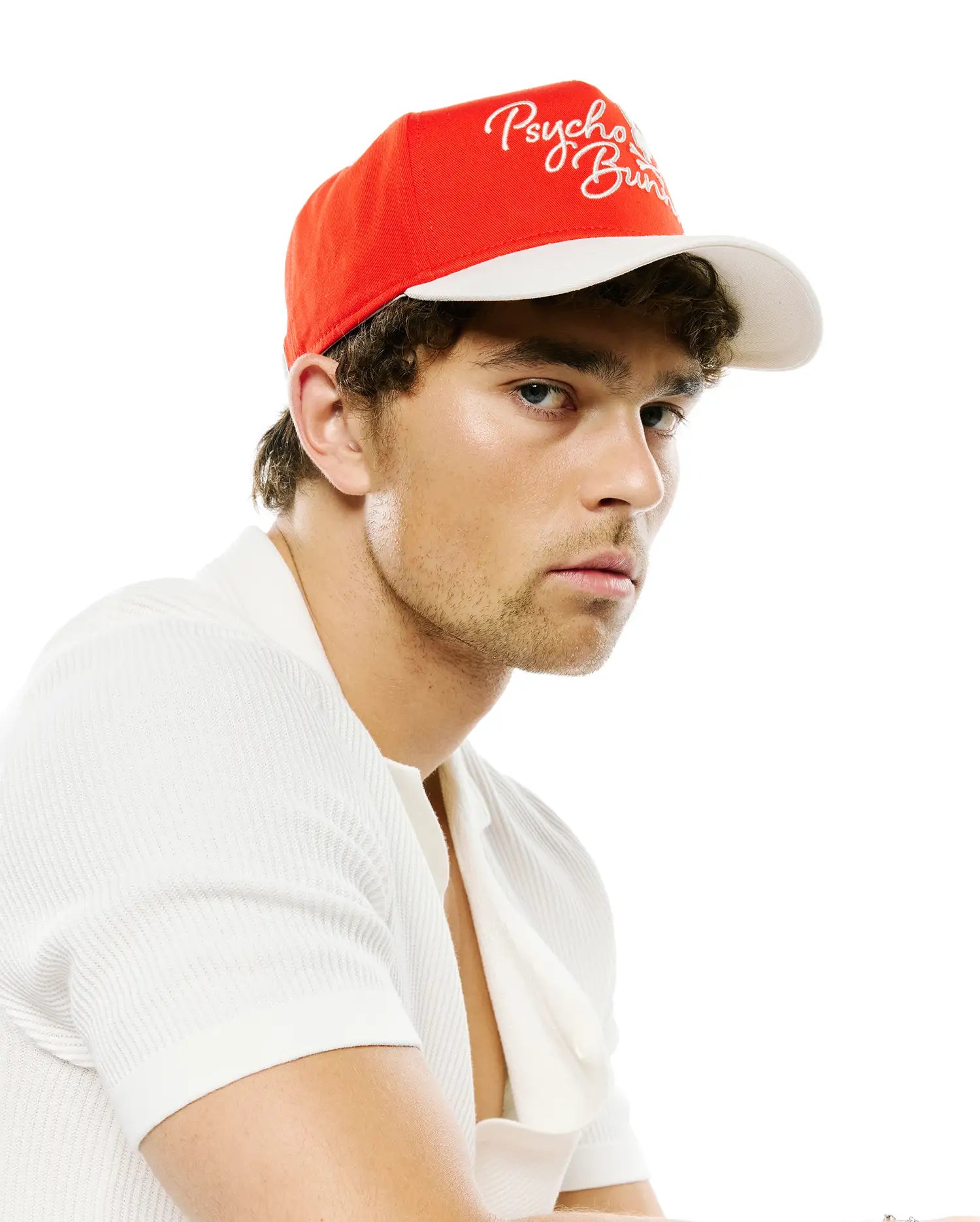 Mens Sable A-Frame Trucker Cap, red with white brim and white embroidered Psycho Bunny script logo, paired with an off-white ribbed knit short-sleeve polo shirt with an open collar.