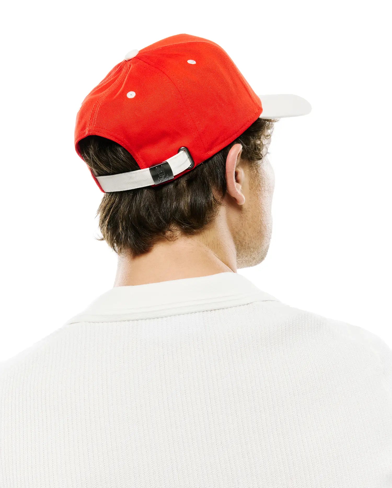 Mens Sable A-Frame Trucker Cap in vibrant red with a white brim, white eyelets, and an adjustable white strap featuring a small black Psycho Bunny skull logo buckle. A white pique knit polo shirt is also visible.