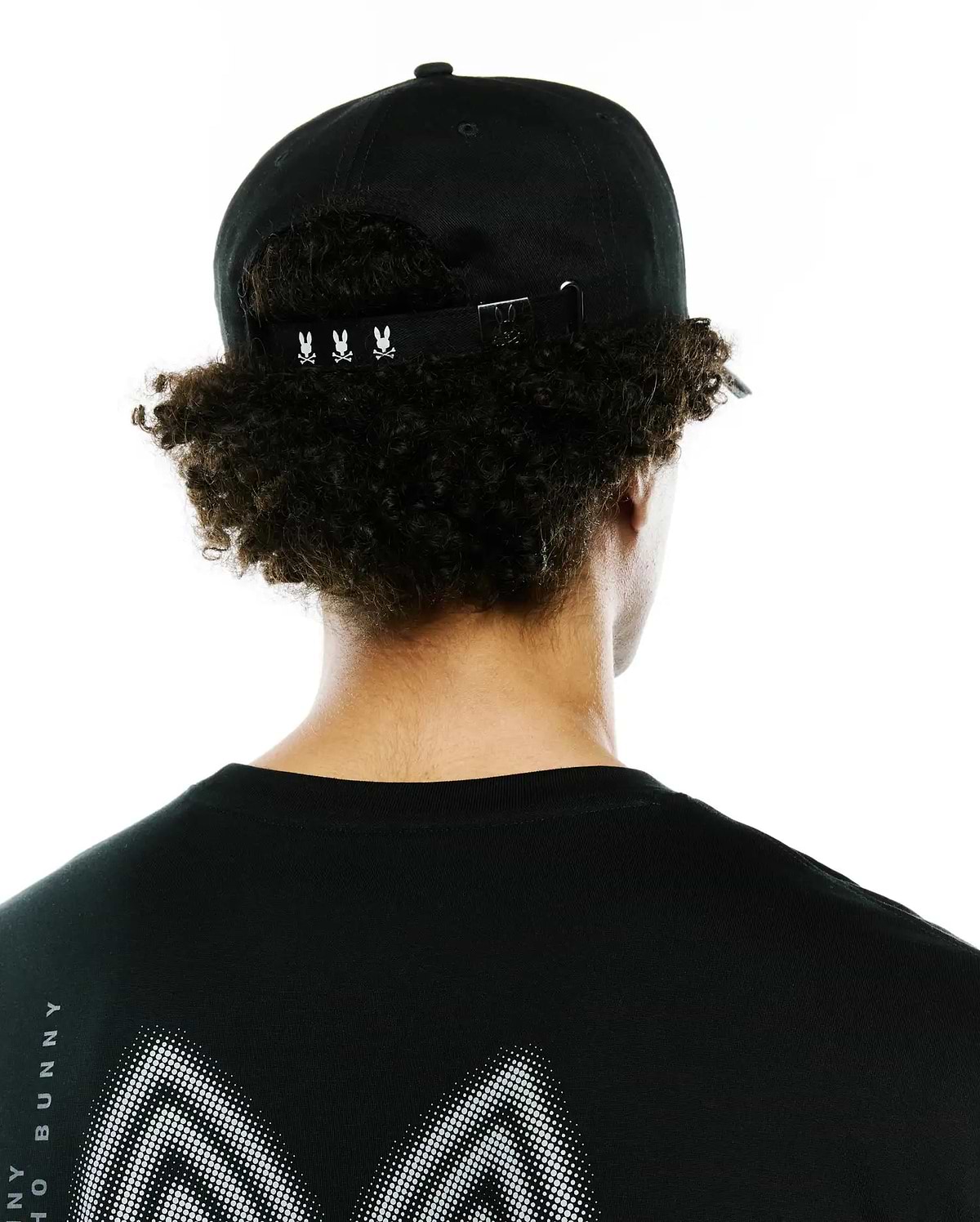 Black Mens Alex A-Frame Trucker Cap with white Psycho Bunny skull logos on the adjustable back strap, paired with a black jersey t-shirt featuring a large white dotted graphic and vertical Psycho Bunny text on the back.
