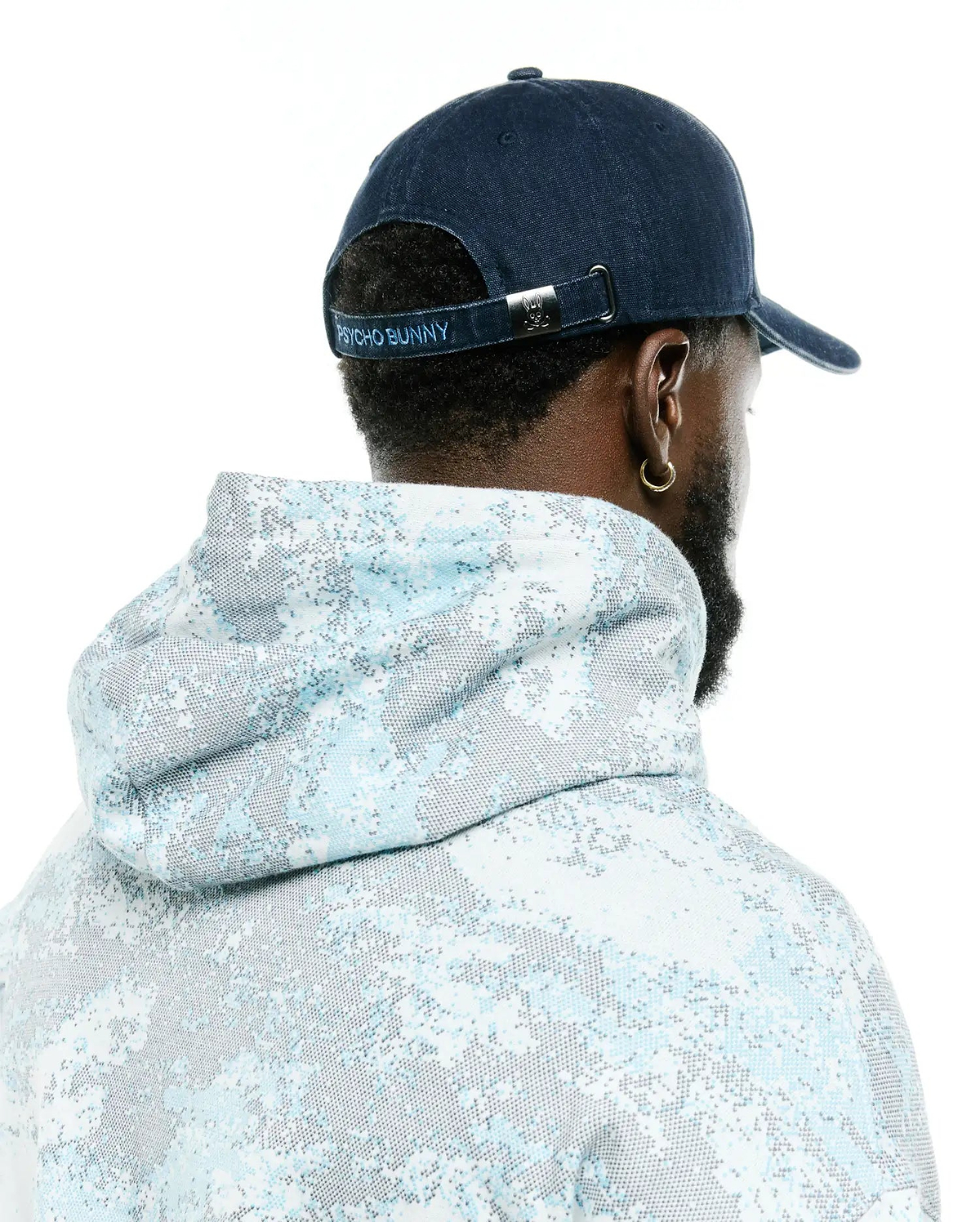 Men's Grover Baseball Cap in navy blue with light blue Psycho Bunny embroidery on the back strap and a silver bunny skull buckle, paired with a relaxed fit hoodie featuring a light blue and grey pixelated all-over print. (3)