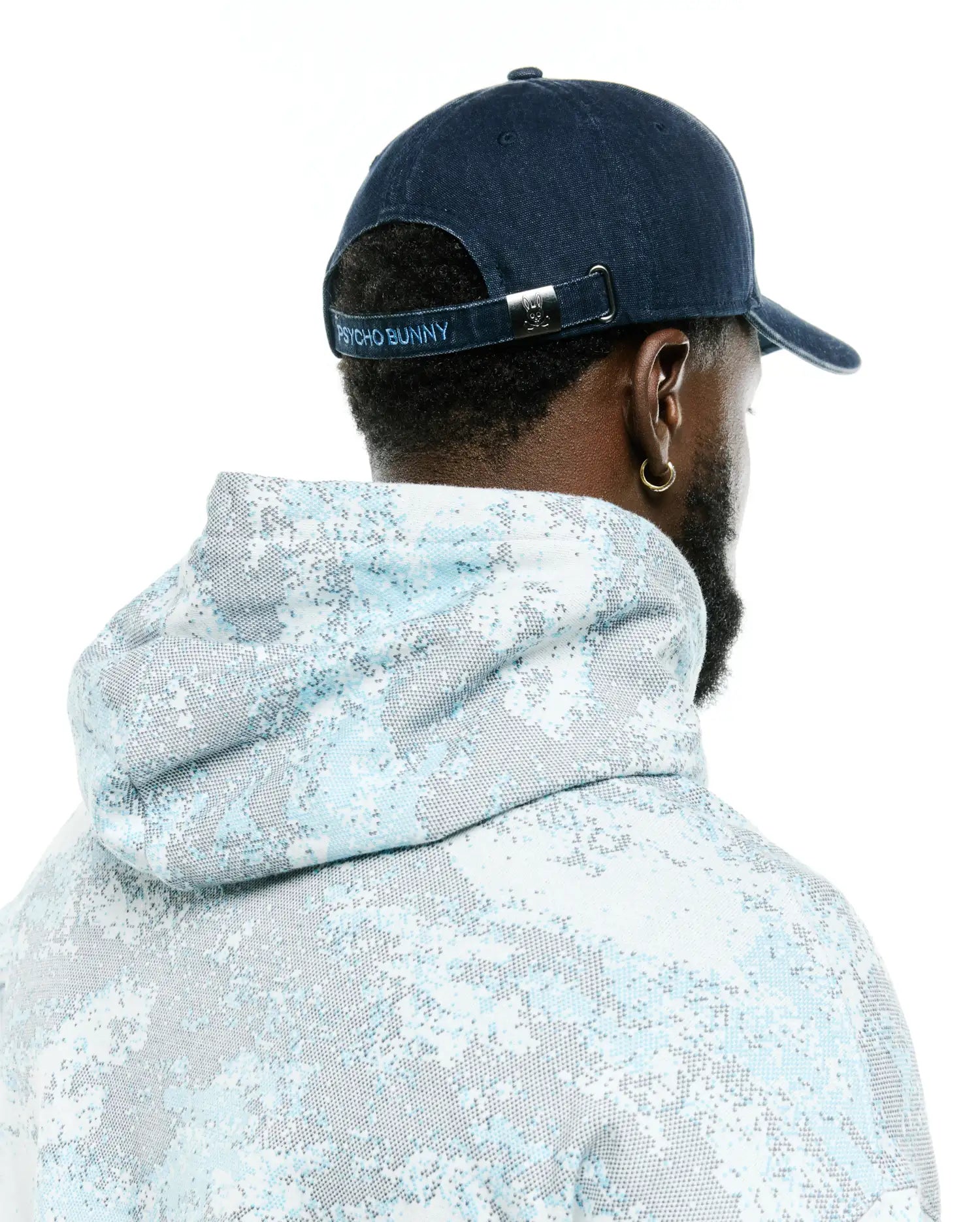 Men's Grover Baseball Cap in navy blue with light blue Psycho Bunny embroidery on the back strap and a silver bunny skull buckle, paired with a relaxed fit hoodie featuring a light blue and grey pixelated all-over print. (3)