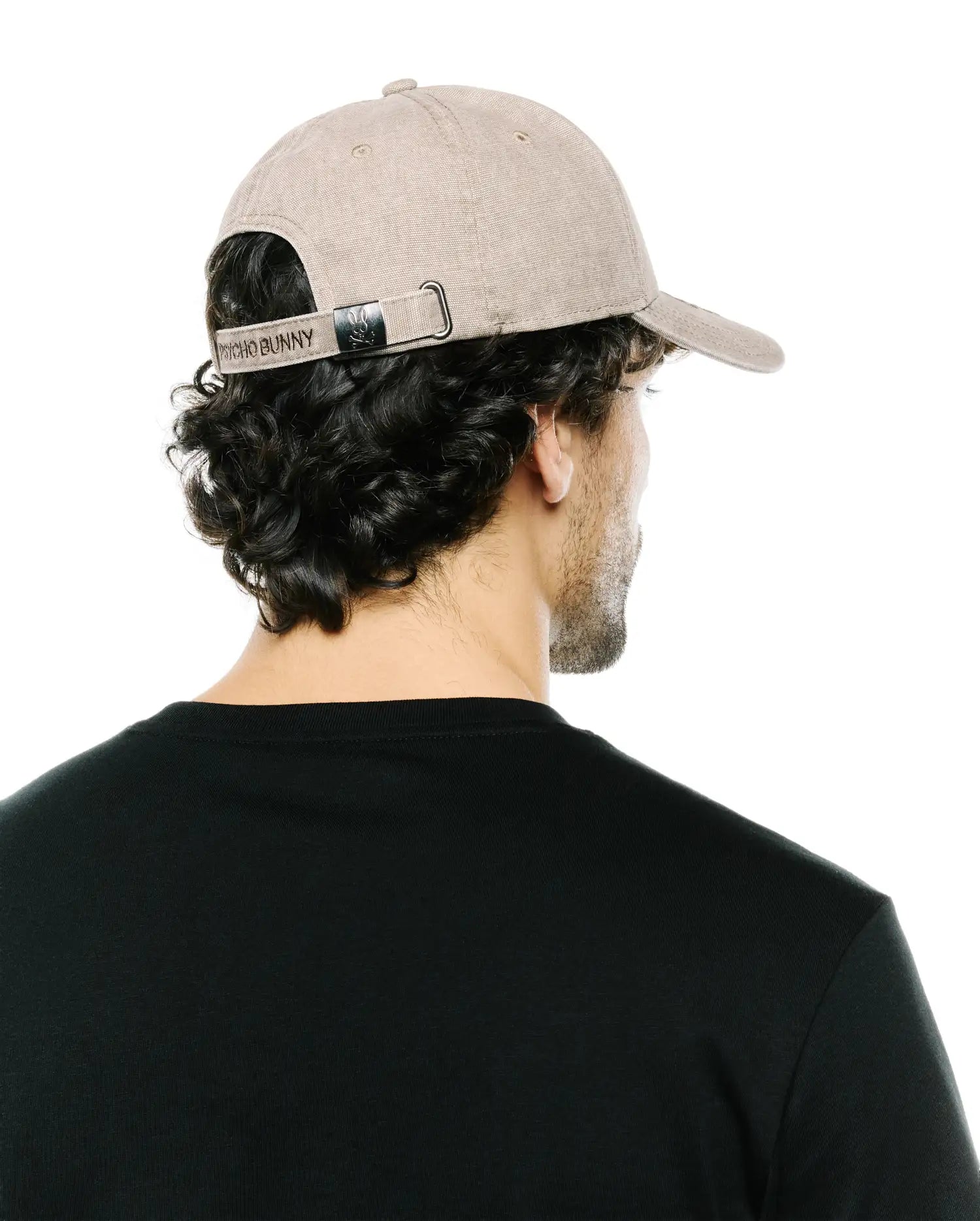 Men's Grover Baseball Cap in beige textured twill, featuring an adjustable back strap with embroidered Psycho Bunny text and a small bunny skull logo on the metallic buckle. (3)