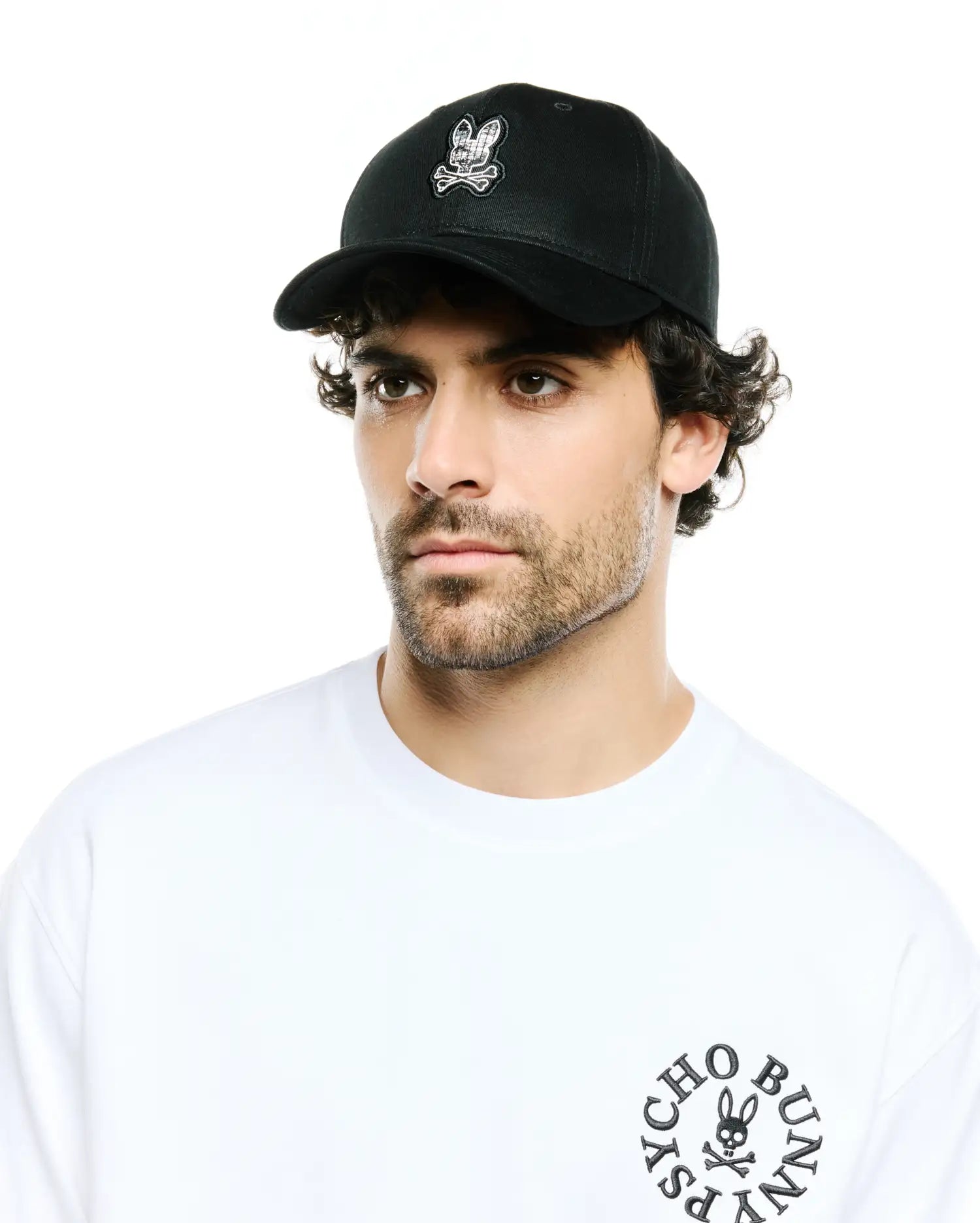 Men's Alfie Baseball Cap in black with a front embroidered white and black plaid bunny skull logo, paired with a white jersey t-shirt featuring a black circular Psycho Bunny text and skull logo on the left chest. (1)