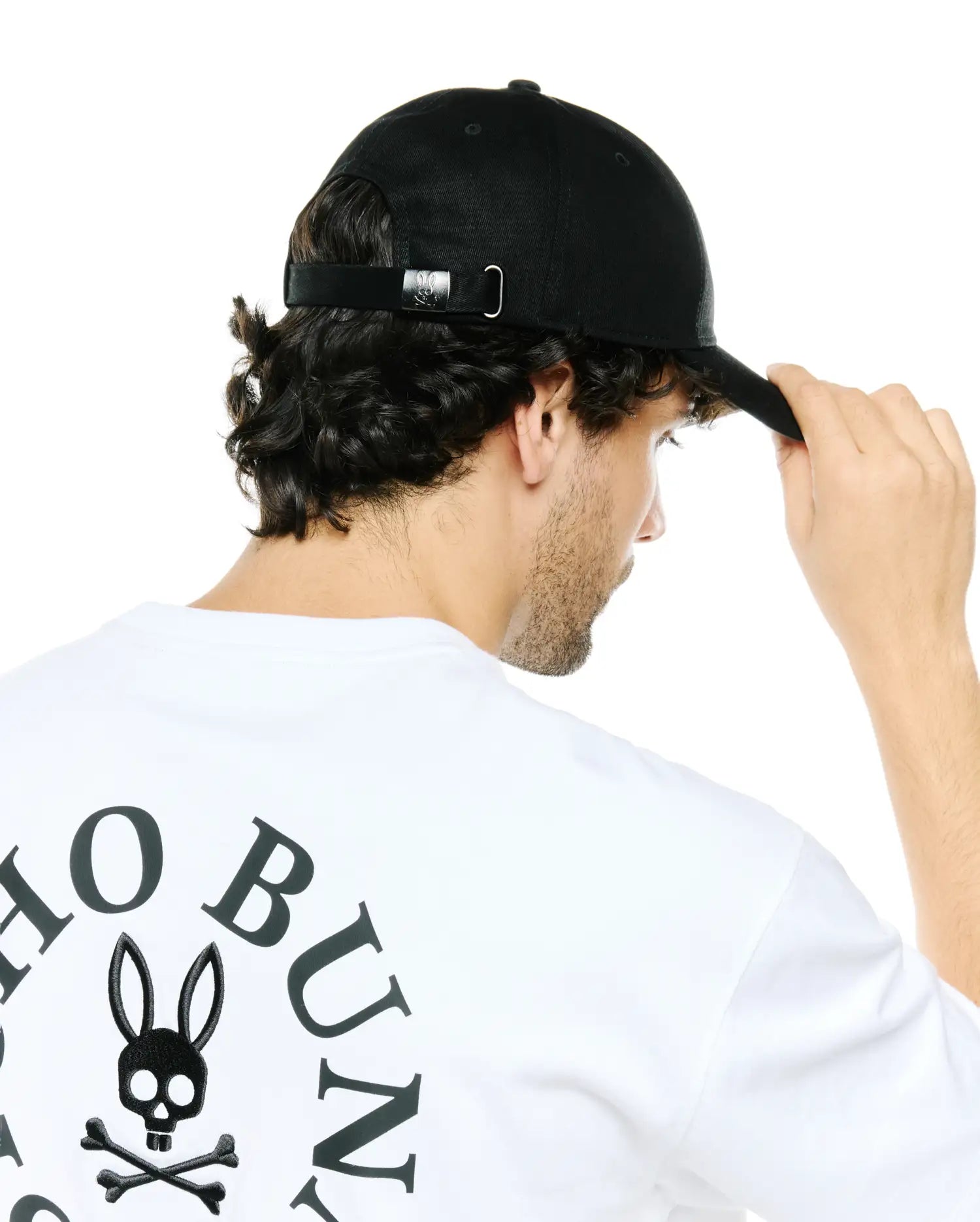 White jersey t-shirt with a large black Psycho Bunny skull and crossbones logo and arched text on the back, paired with a black Men's Alfie Baseball Cap featuring a metallic bunny logo on the adjustable strap. (3)