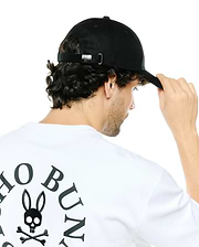 White jersey t-shirt with a large black Psycho Bunny skull and crossbones logo and arched text on the back, paired with a black Men's Alfie Baseball Cap featuring a metallic bunny logo on the adjustable strap. (3)