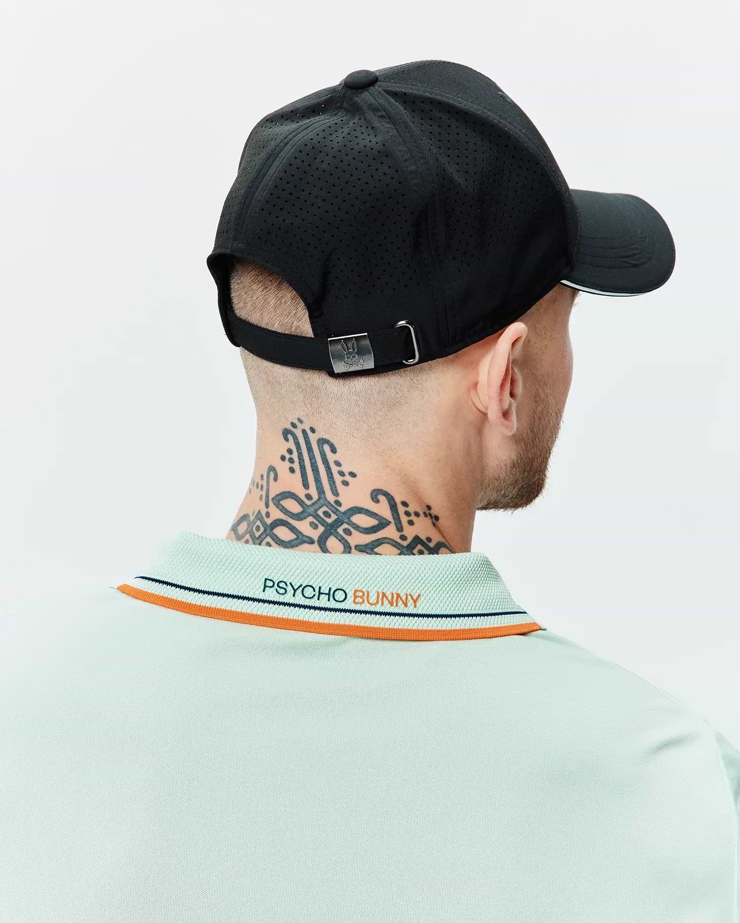Mens Blaze Sport Cap in black with perforated texture, white brim stripe, and metal bunny skull buckle, paired with a light green pique polo featuring orange and navy collar stripes and embroidered Psycho Bunny text on the back.