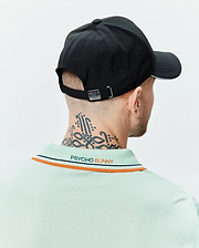 Mens Blaze Sport Cap in black with perforated texture, white brim stripe, and metal bunny skull buckle, paired with a light green pique polo featuring orange and navy collar stripes and embroidered Psycho Bunny text on the back.