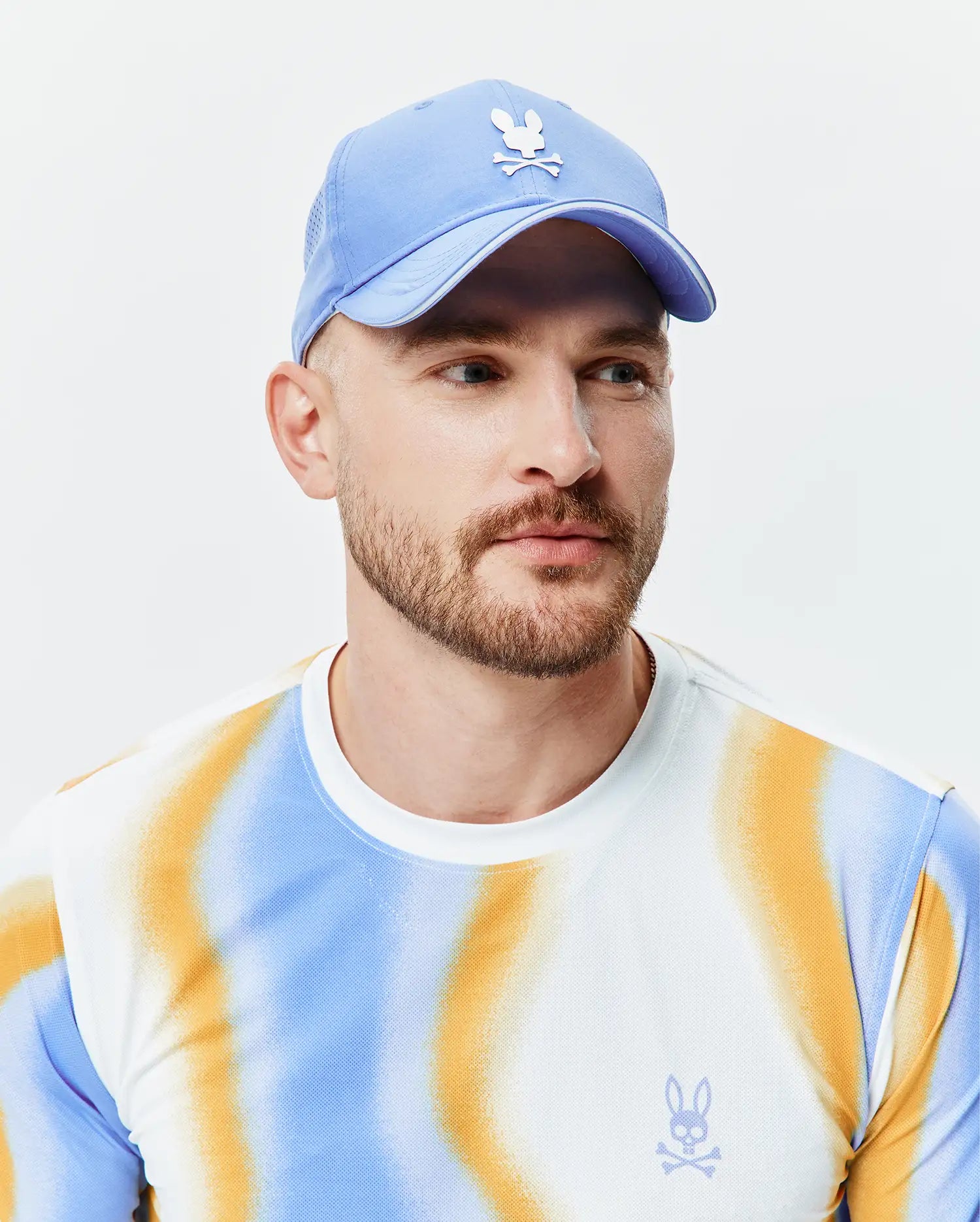 Mens Blaze Sport Cap, a light blue mesh cap with white embroidered Psycho Bunny logo. White pique long-sleeve t-shirt with blue/orange gradient stripes and small blue embroidered Psycho Bunny chest logo. (1)