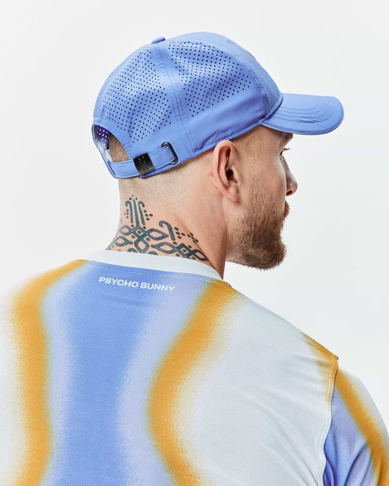 Mens Blaze Sport Cap in periwinkle blue with perforated panels and a dark metal buckle, paired with a white mesh t-shirt, gradient blue and orange pattern, and Psycho Bunny text on the back. (3)