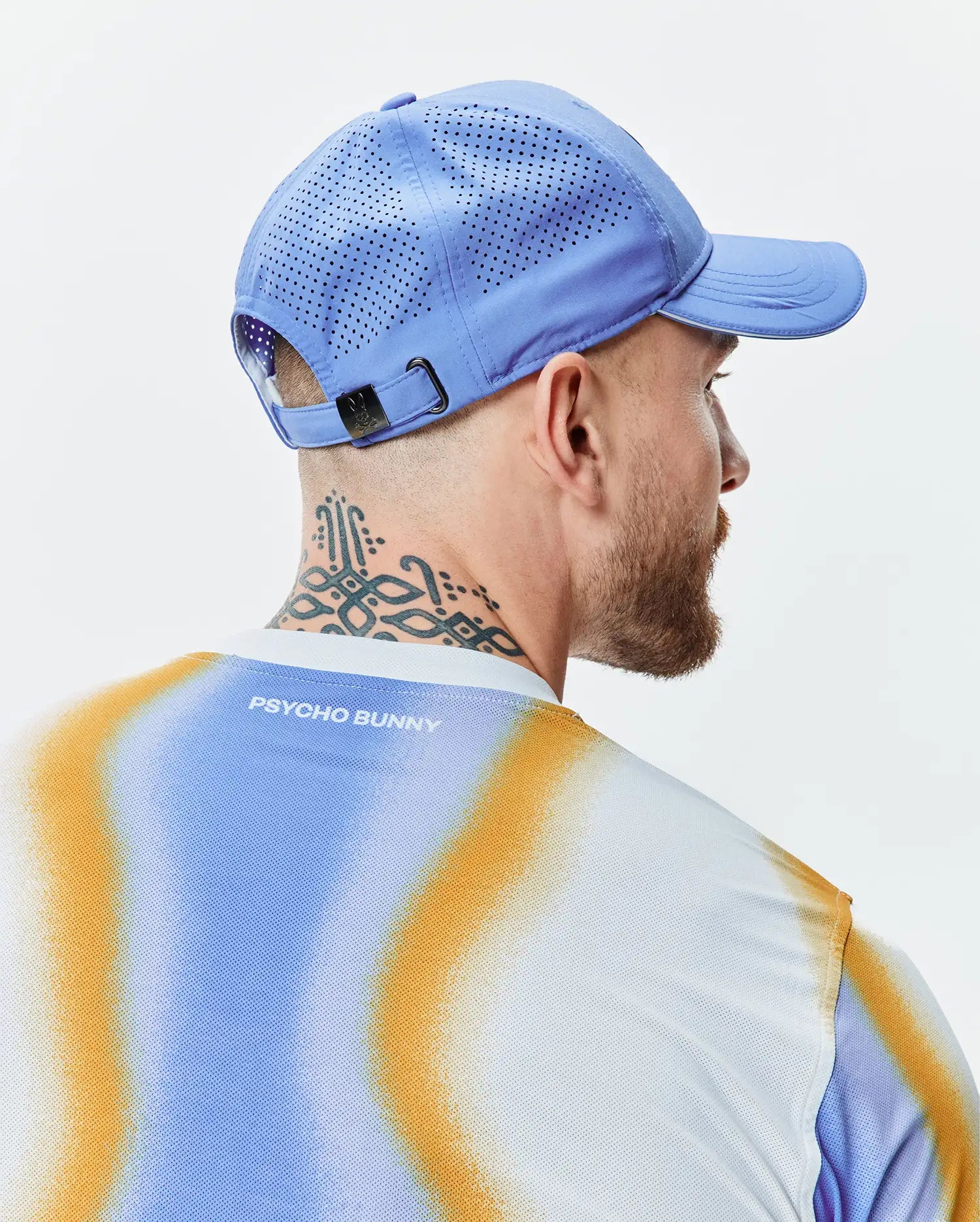 Mens Blaze Sport Cap in periwinkle blue with perforated panels and a dark metal buckle, paired with a white mesh t-shirt, gradient blue and orange pattern, and Psycho Bunny text on the back. (3)