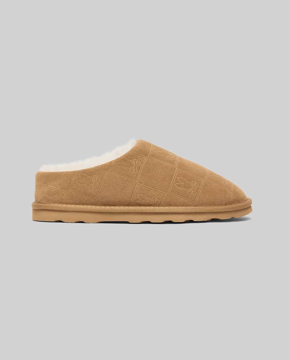 Mens Gio Suede Shearling Slippers in tan suede with plush white shearling lining, featuring an all-over debossed Psycho Bunny skull logo in a diamond grid pattern.