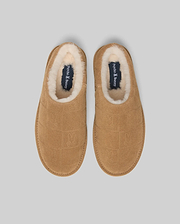 Mens Gio Suede Shearling Slippers in tan suede, featuring an all-over embossed Psycho Bunny skull logo pattern, plush cream shearling lining, and a navy insole with the Psycho Bunny text logo.