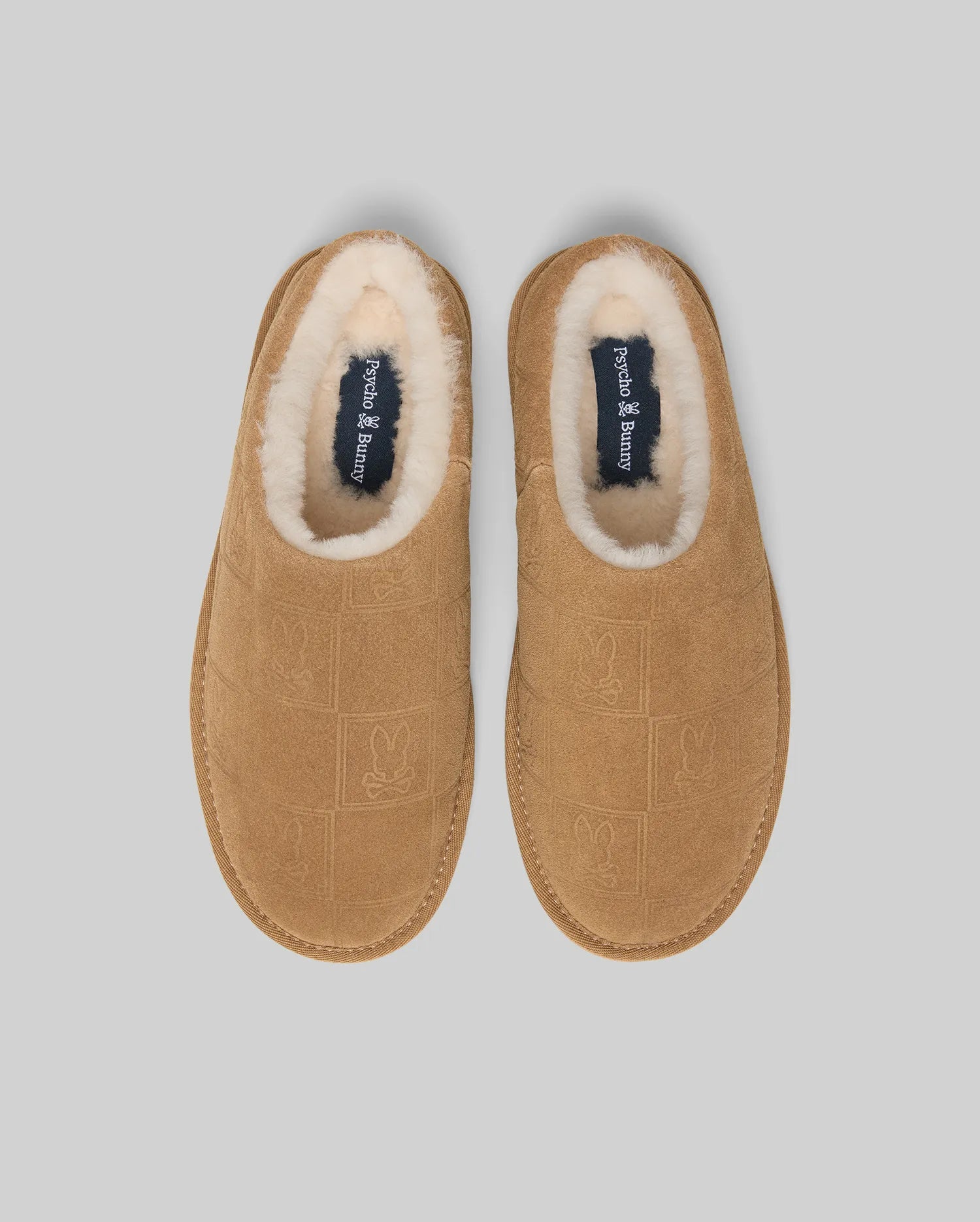 Mens Gio Suede Shearling Slippers in tan suede, featuring an all-over embossed Psycho Bunny skull logo pattern, plush cream shearling lining, and a navy insole with the Psycho Bunny text logo.