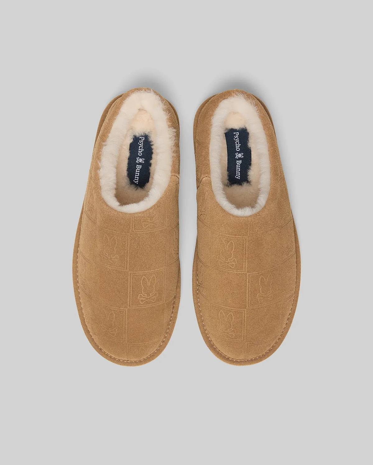 Mens Gio Suede Shearling Slippers in tan suede, featuring an all-over embossed Psycho Bunny skull logo pattern, plush cream shearling lining, and a navy insole with the Psycho Bunny text logo.