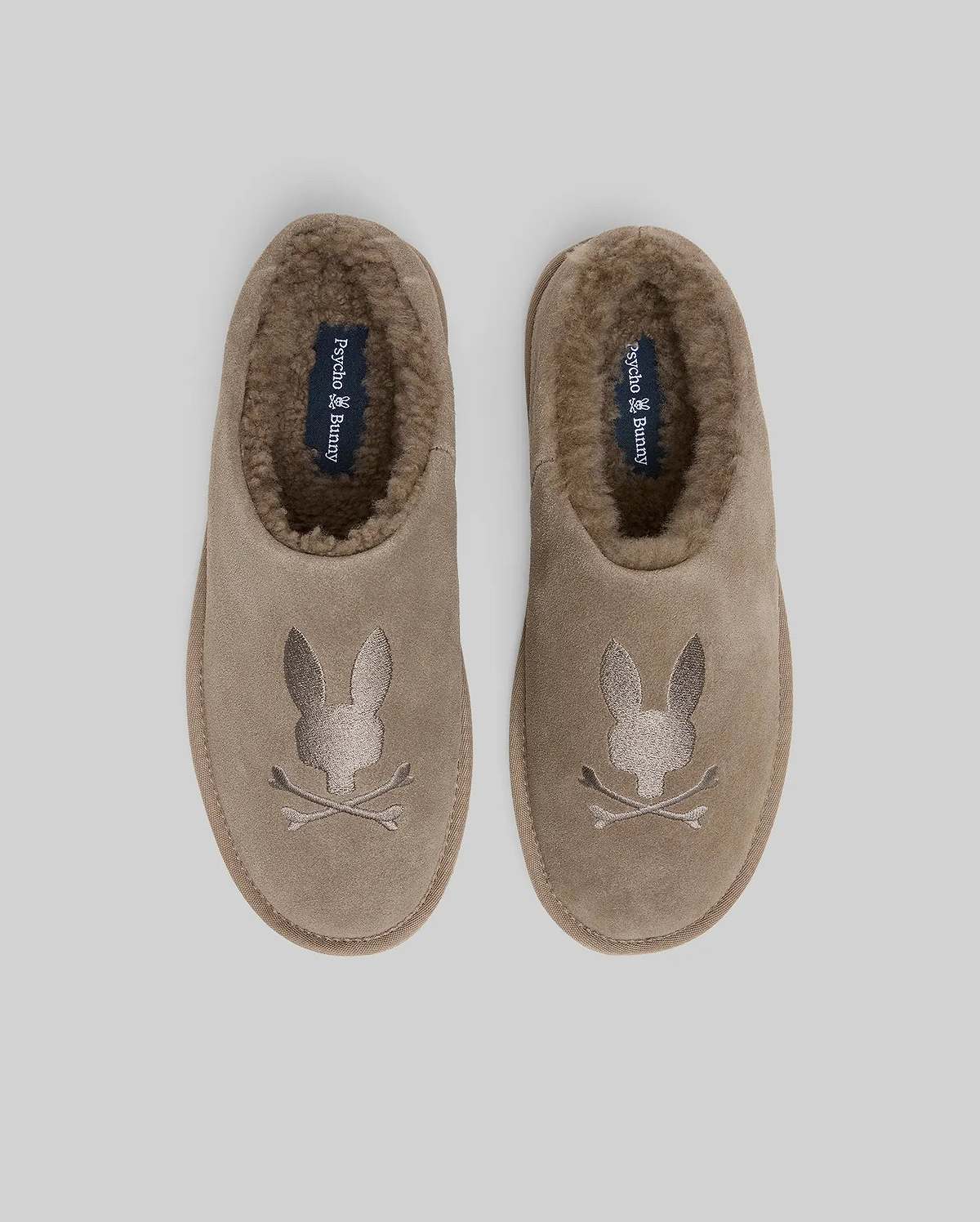 Mens Suede Sherpa Bunny Slipper in taupe, featuring a plush brown sherpa lining and a large embroidered Psycho Bunny skull logo on the toe, with brand text on the insole.