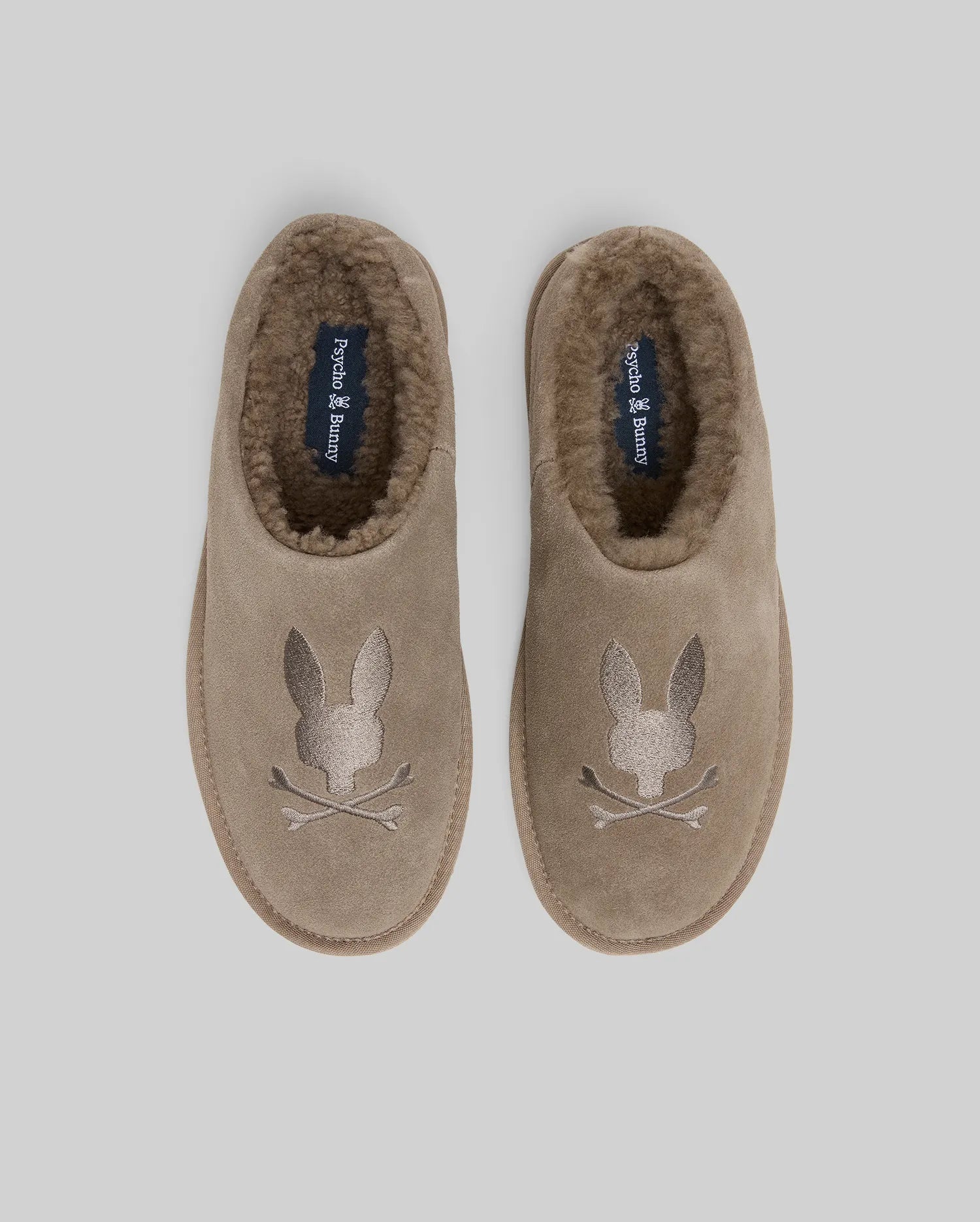Mens Suede Sherpa Bunny Slipper in taupe, featuring a plush brown sherpa lining and a large embroidered Psycho Bunny skull logo on the toe, with brand text on the insole.