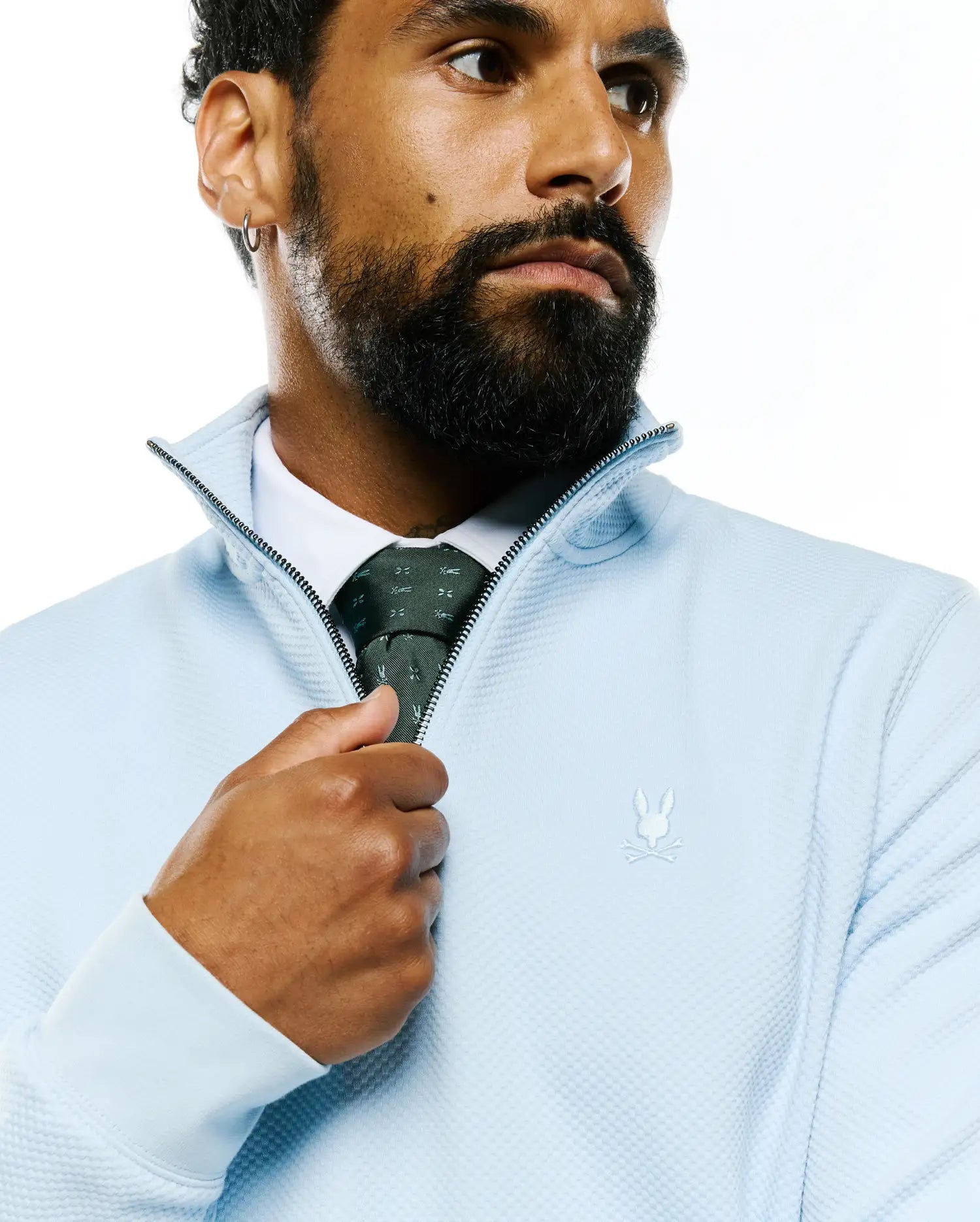 Light blue textured quarter-zip pullover with a tone-on-tone embroidered Psycho Bunny chest logo, paired with a dark green Mens Bunny Neat Woven Tie featuring a subtle bunny pattern.