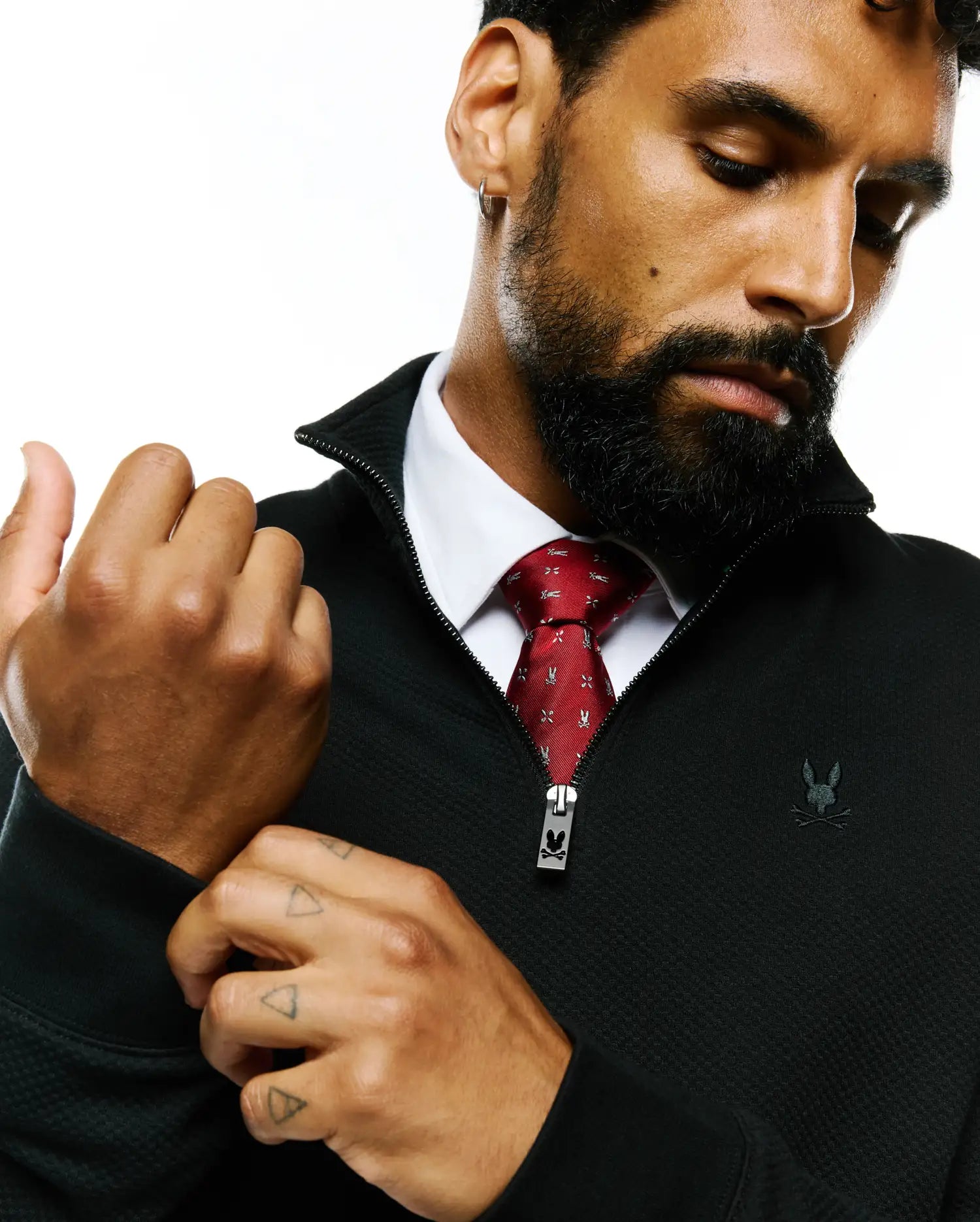 Black textured quarter-zip with embroidered Psycho Bunny skull logo on chest and branded zipper pull, worn over a white shirt and a red Mens Bunny Neat Woven Tie featuring an all-over skull pattern.