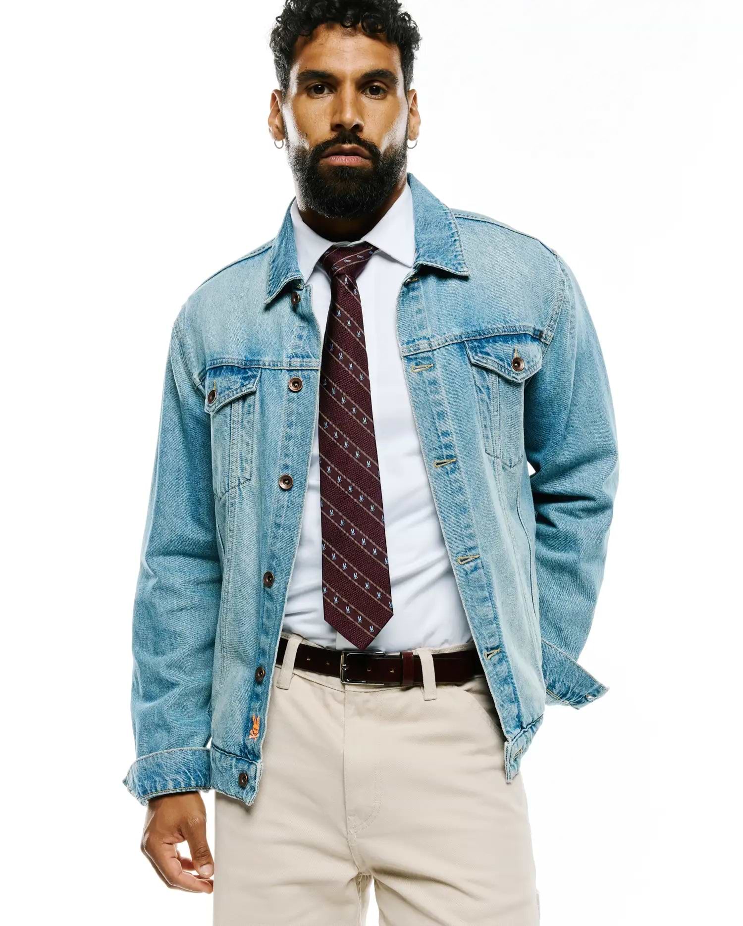 Mens Bunny Stripe Woven Tie in burgundy with white diagonal Psycho Bunny skull logos, paired with a crisp white collared shirt, light wash denim jacket featuring an orange cuff logo, and beige chinos.