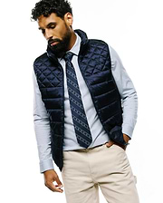 Navy quilted puffer vest with small embroidered Psycho Bunny skull logo, layered over a light blue striped button-down shirt, a MENS BUNNY STRIPE WOVEN TIE, and classic fit khaki chinos.