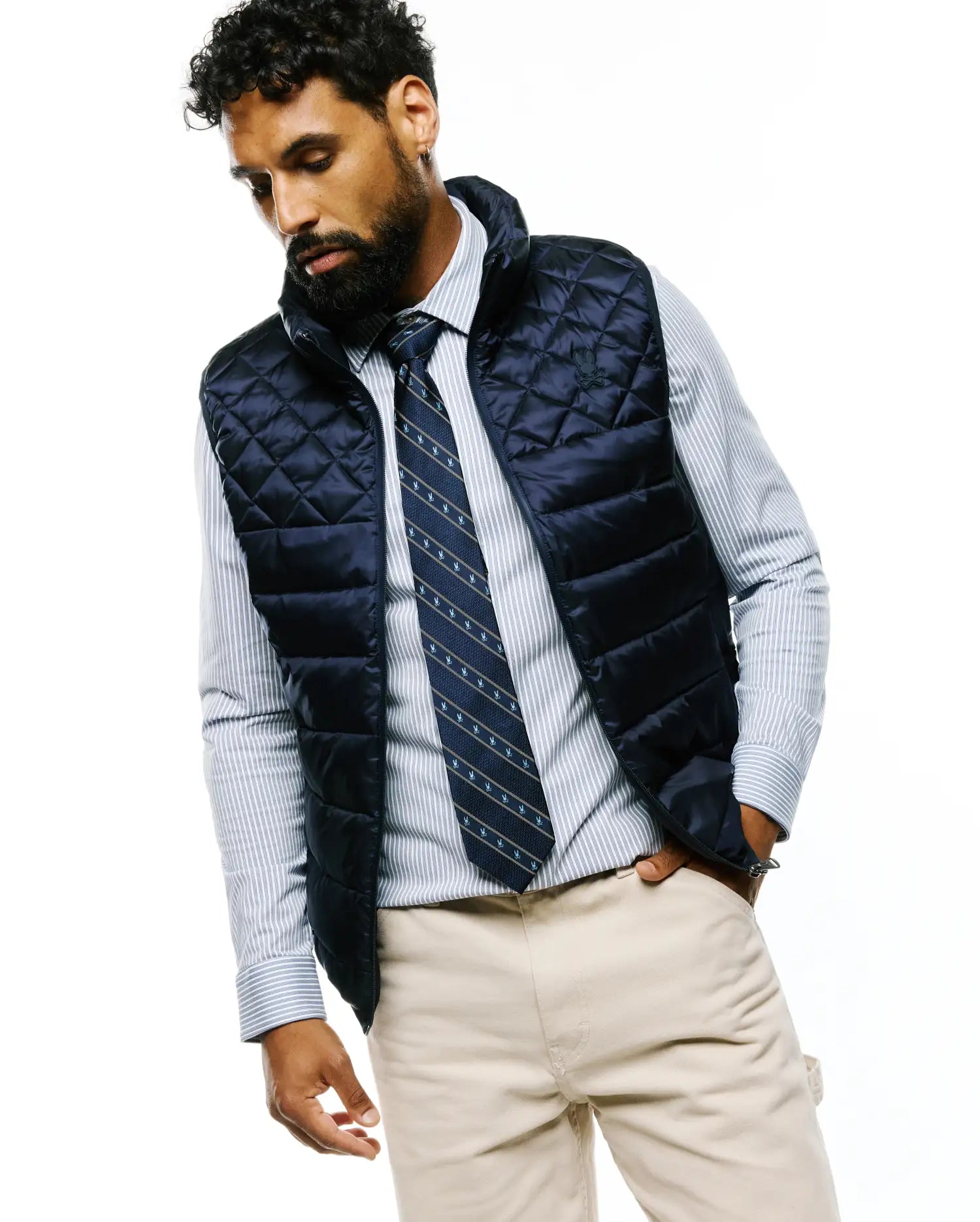 Navy quilted puffer vest with small embroidered Psycho Bunny skull logo, layered over a light blue striped button-down shirt, a MENS BUNNY STRIPE WOVEN TIE, and classic fit khaki chinos.