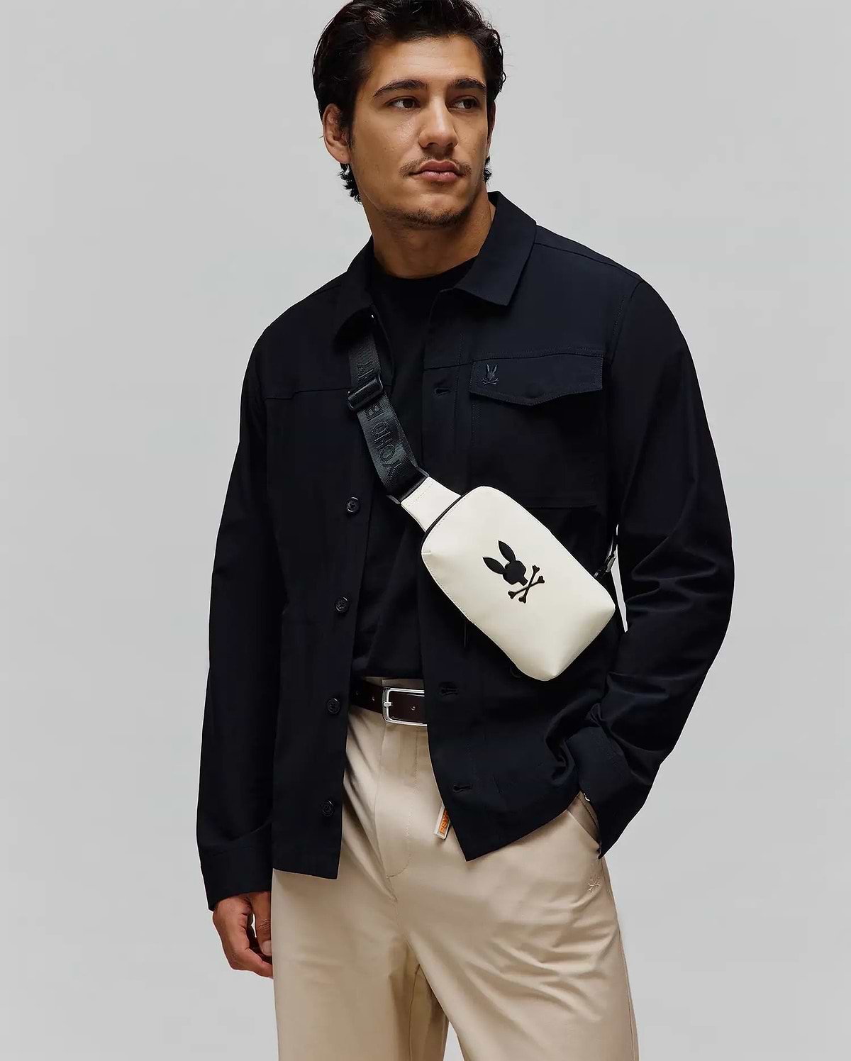 Mens Rubberized Crossbody in cream with a large black Psycho Bunny skull logo, worn with a black collared shirt jacket featuring a small embroidered bunny, a black crew neck tee, and khaki pants.