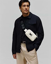 Mens Rubberized Crossbody in cream with a large black Psycho Bunny skull logo, worn with a black collared shirt jacket featuring a small embroidered bunny, a black crew neck tee, and khaki pants.