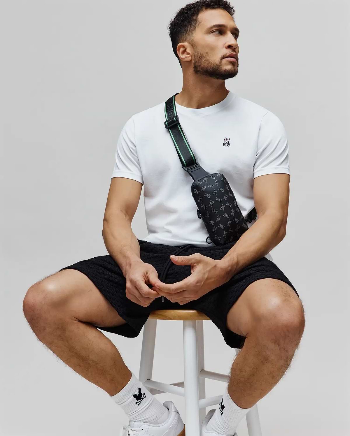 White jersey t-shirt with embroidered pink-eared Psycho Bunny skull, black textured shorts, and a black Mens Rubberized Crossbody bag with all-over bunny skull print and green-striped strap.