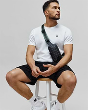 White jersey t-shirt with embroidered pink-eared Psycho Bunny skull, black textured shorts, and a black Mens Rubberized Crossbody bag with all-over bunny skull print and green-striped strap.