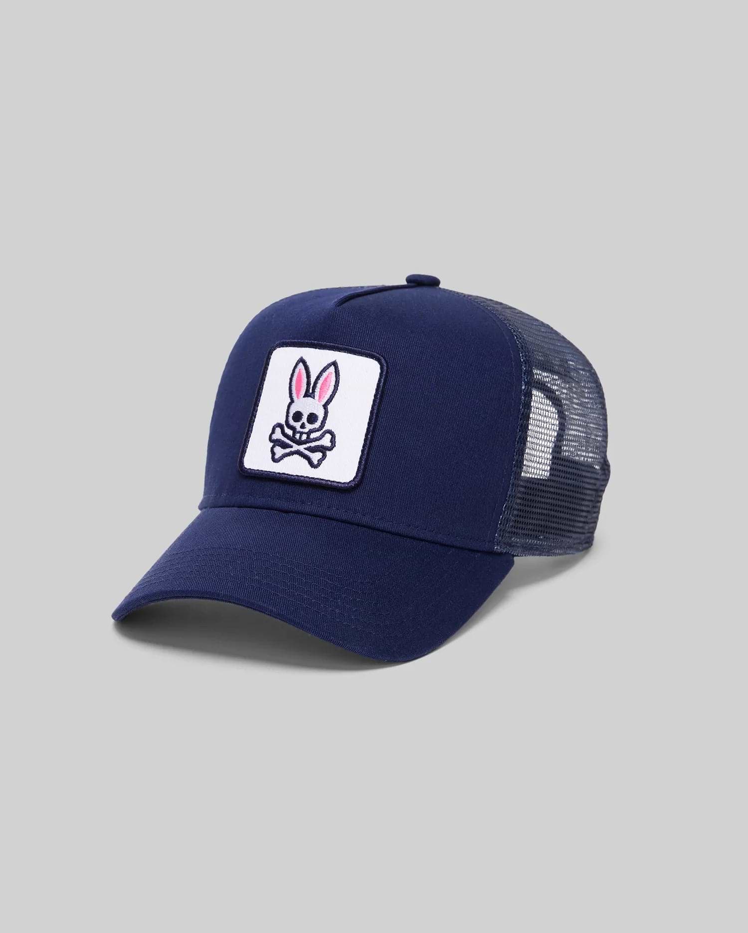 The Psycho Bunny MENS BOLD DAD TRUCKER CAP (MO2600001) is a navy blue cap with a curved brim, mesh sides and back, and features a front patch showing a pink-eared bunny above crossed bones on a white background. (1)