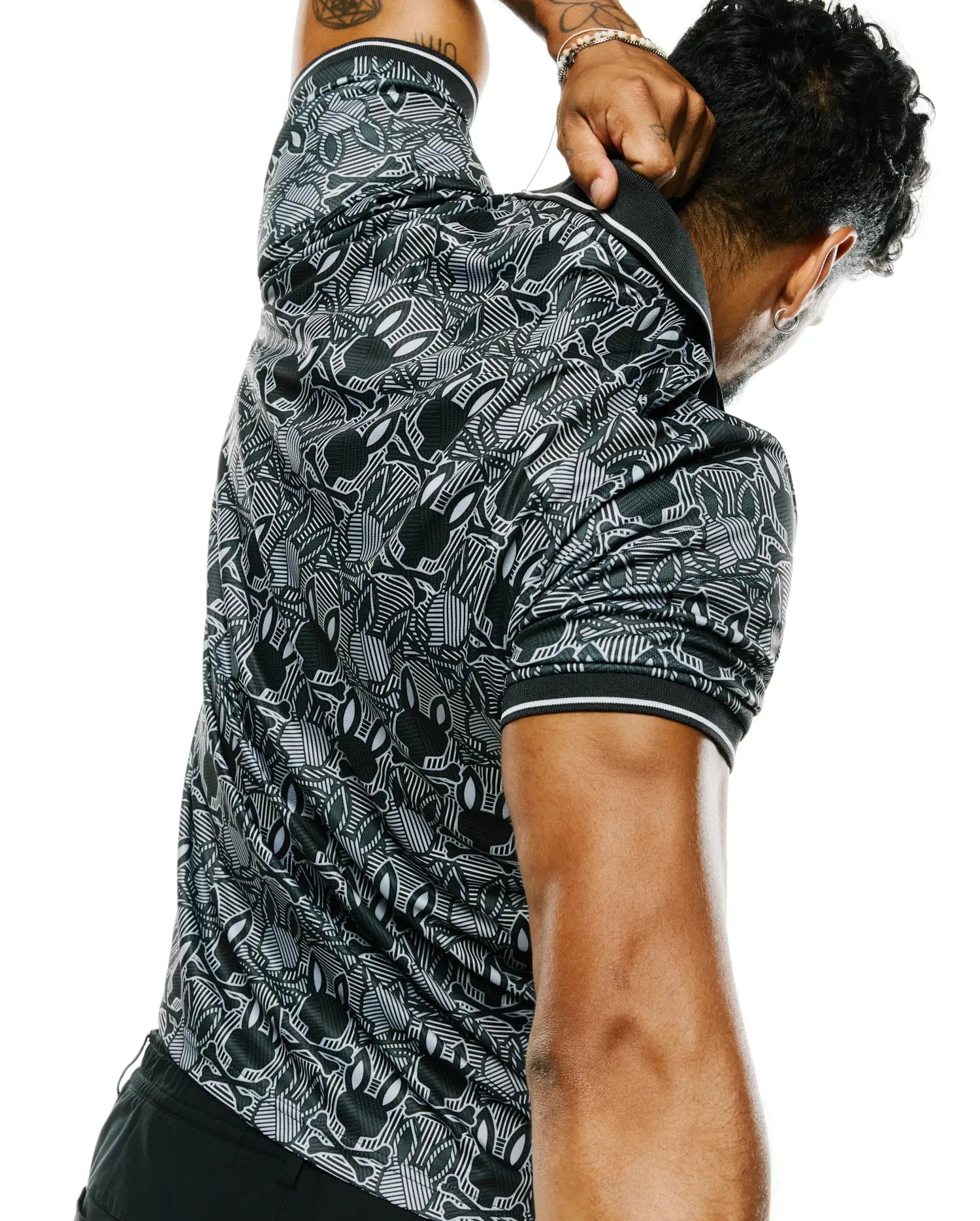 Mens Reeves All Over Bunny Sport Polo, a black and white performance jersey polo with a classic fit, featuring an intricate all-over Psycho Bunny skull and geometric pattern, with white striped collar and sleeve cuffs.