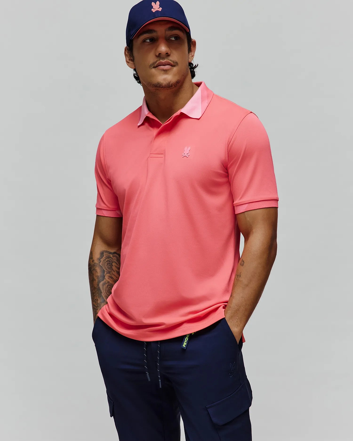 Mens Onyx Sport Polo, a vibrant coral performance polo with a classic fit, features a lighter pink collar underside, ribbed cuffs, and a small tonal embroidered Psycho Bunny skull logo on the left chest.