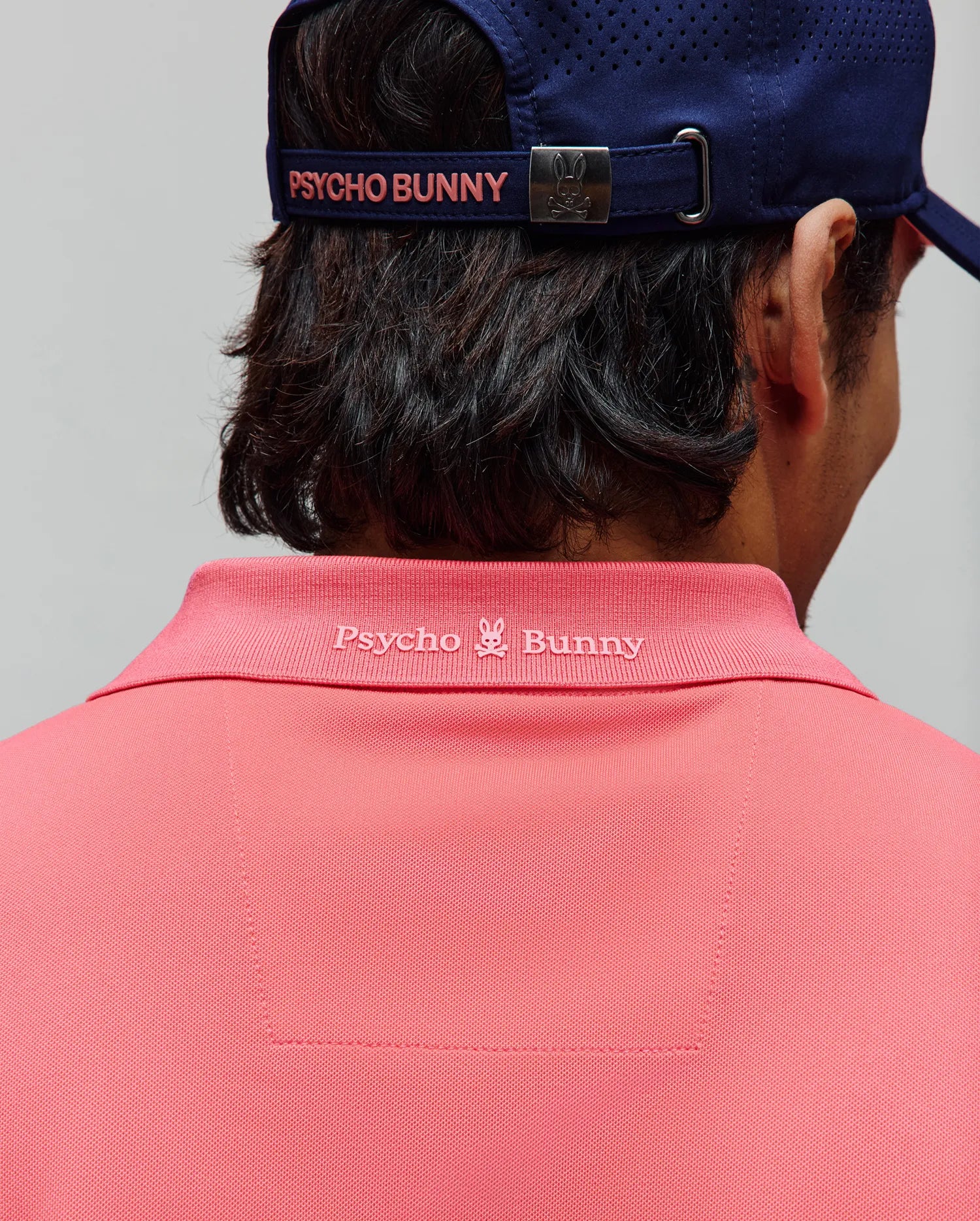 Mens Onyx Sport Polo in vibrant coral pink pique knit, featuring a tonal embroidered Psycho Bunny logo and text on the back collar, complemented by a navy perforated cap with pink branding.