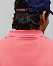 Mens Onyx Sport Polo in vibrant coral pink pique knit, featuring a tonal embroidered Psycho Bunny logo and text on the back collar, complemented by a navy perforated cap with pink branding.