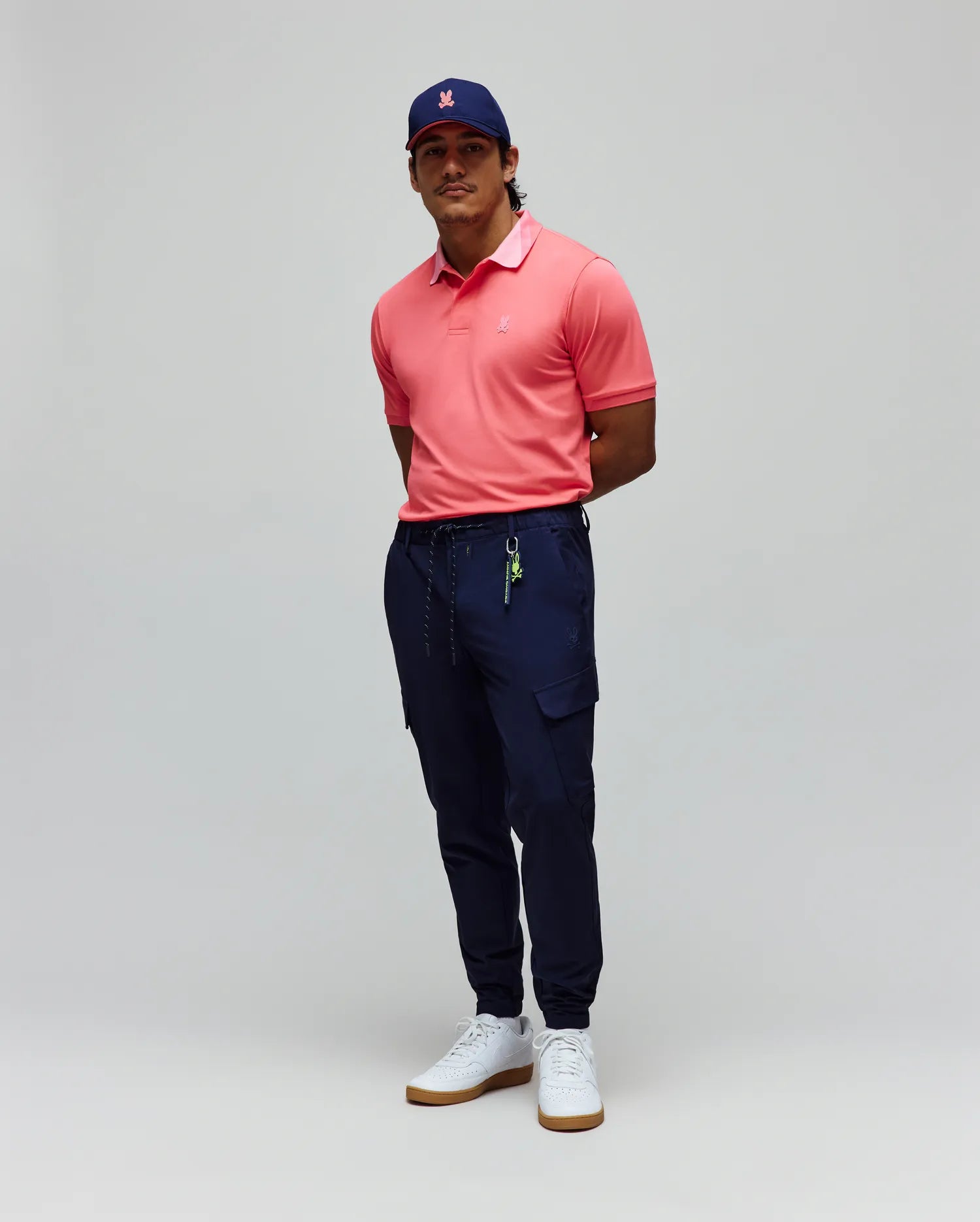 Mens Onyx Sport Polo in coral pique with a lighter pink collar and embroidered chest logo, paired with navy slim-fit cargo joggers featuring a tonal bunny logo and a navy cap with a pink embroidered bunny.