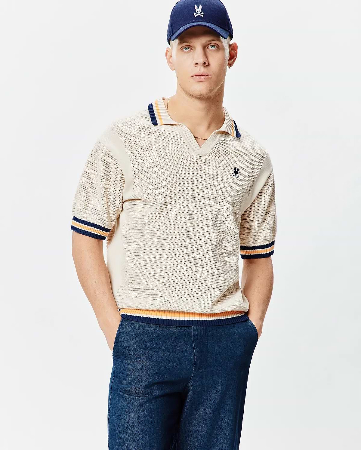Mens Theros Knit Polo, a cream open-knit polo with a relaxed fit, V-neck, and navy, orange, yellow striped ribbed collar, cuffs, and hem, featuring a small embroidered Psycho Bunny skull logo on the left chest.