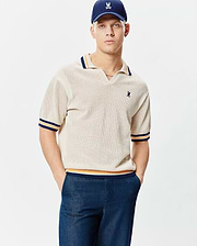 Mens Theros Knit Polo, a cream open-knit polo with a relaxed fit, V-neck, and navy, orange, yellow striped ribbed collar, cuffs, and hem, featuring a small embroidered Psycho Bunny skull logo on the left chest.