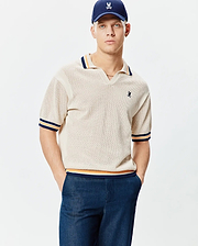 Mens Theros Knit Polo, a cream open-knit polo with a relaxed fit, V-neck, and navy, orange, yellow striped ribbed collar, cuffs, and hem, featuring a small embroidered Psycho Bunny skull logo on the left chest.