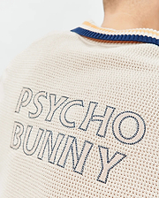 Mens Theros Knit Polo in cream with a textured open knit, featuring a navy and orange striped collar and large embroidered PSYCHO BUNNY text in navy and light beige on the upper back.