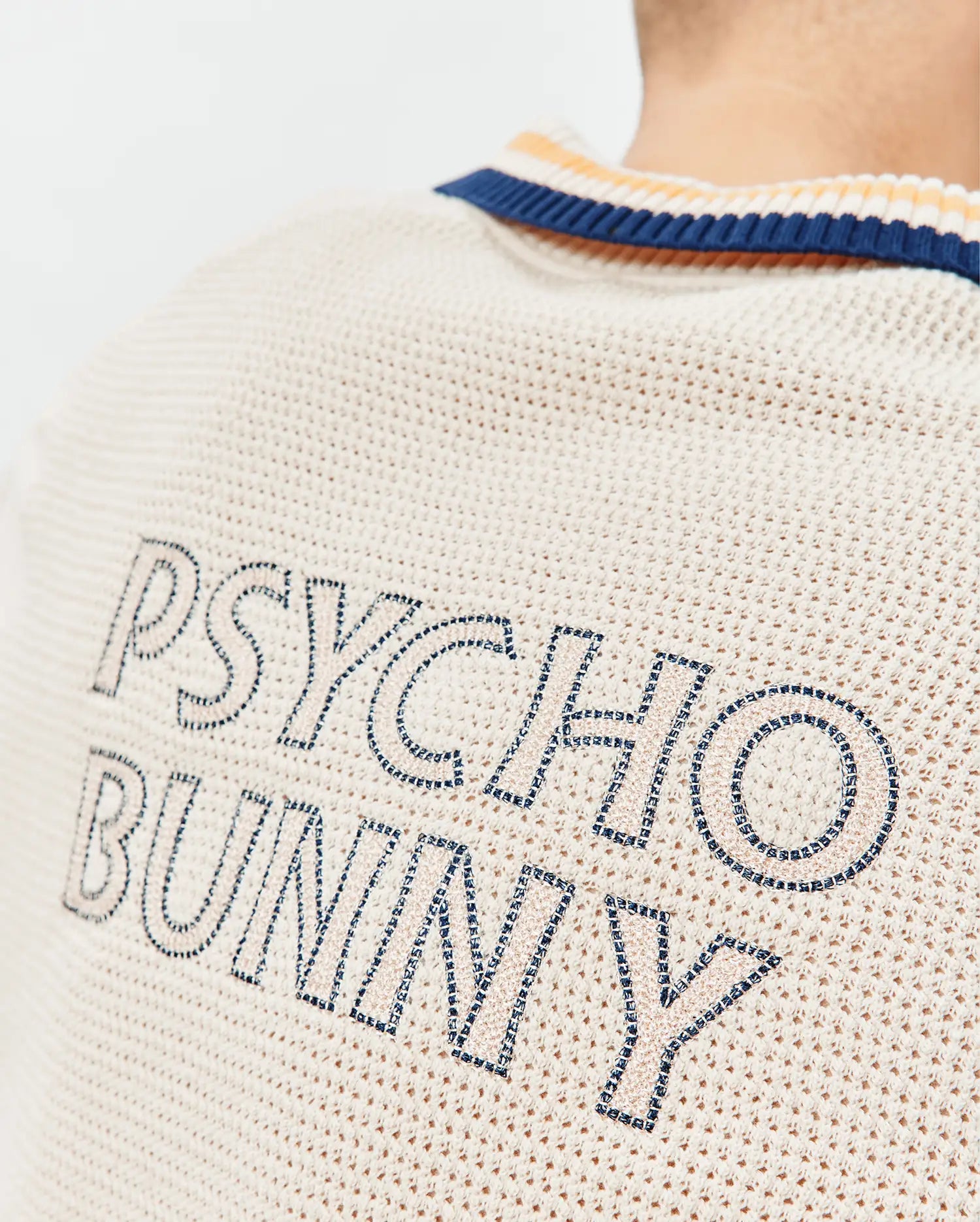 Mens Theros Knit Polo in cream with a textured open knit, featuring a navy and orange striped collar and large embroidered PSYCHO BUNNY text in navy and light beige on the upper back.
