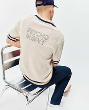 Cream Mens Theros Knit Polo with a relaxed fit, featuring an open knit texture, navy and orange striped ribbed collar, cuffs, and hem, and a large outlined PSYCHO BUNNY text logo on the upper back.