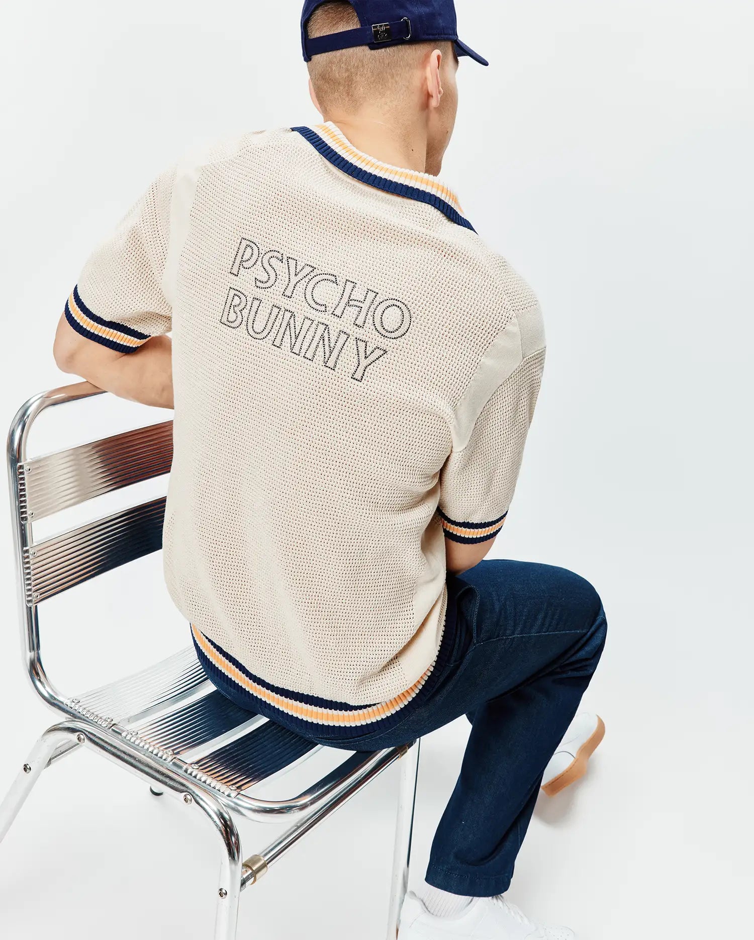 Cream Mens Theros Knit Polo with a relaxed fit, featuring an open knit texture, navy and orange striped ribbed collar, cuffs, and hem, and a large outlined PSYCHO BUNNY text logo on the upper back.