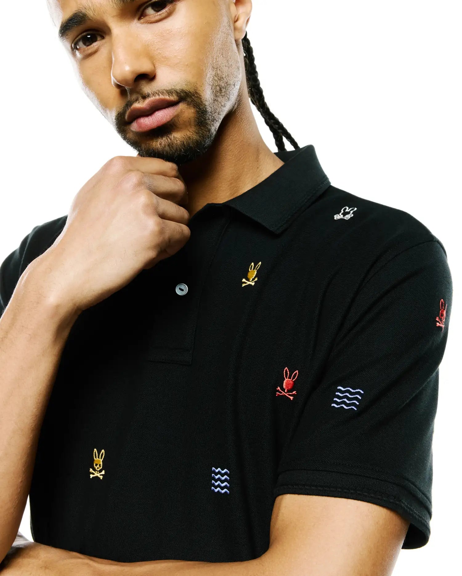 Mens Colin All-Over Pique Polo, black with classic fit, featuring all-over embroidered Psycho Bunny skull logos in gold, red, and white, plus blue wave patterns.