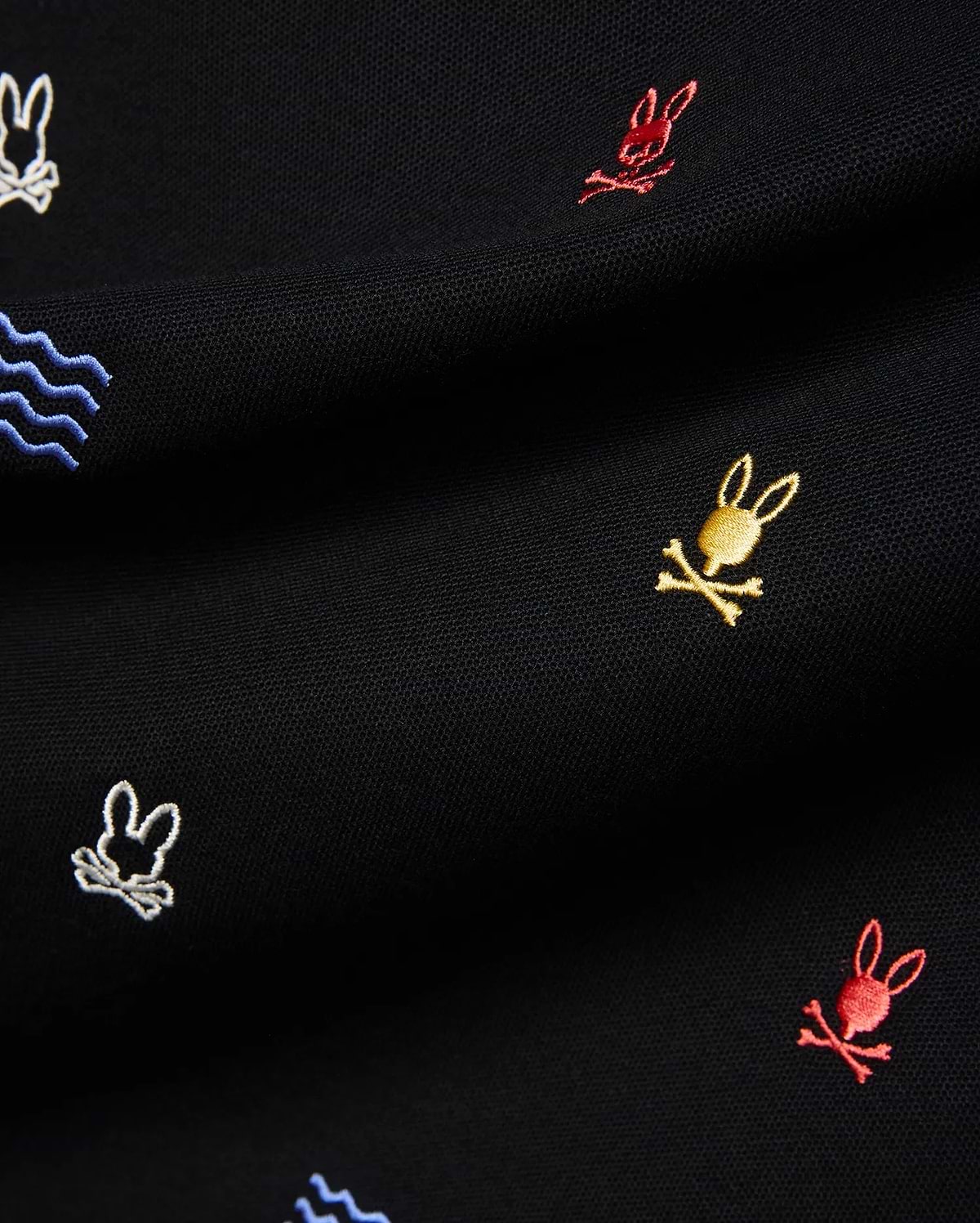 Mens Colin All-Over Pique Polo in black, featuring a classic pique texture with an all-over embroidered pattern of small, colorful Psycho Bunny skull logos in red, yellow, and white, alongside blue wave motifs.