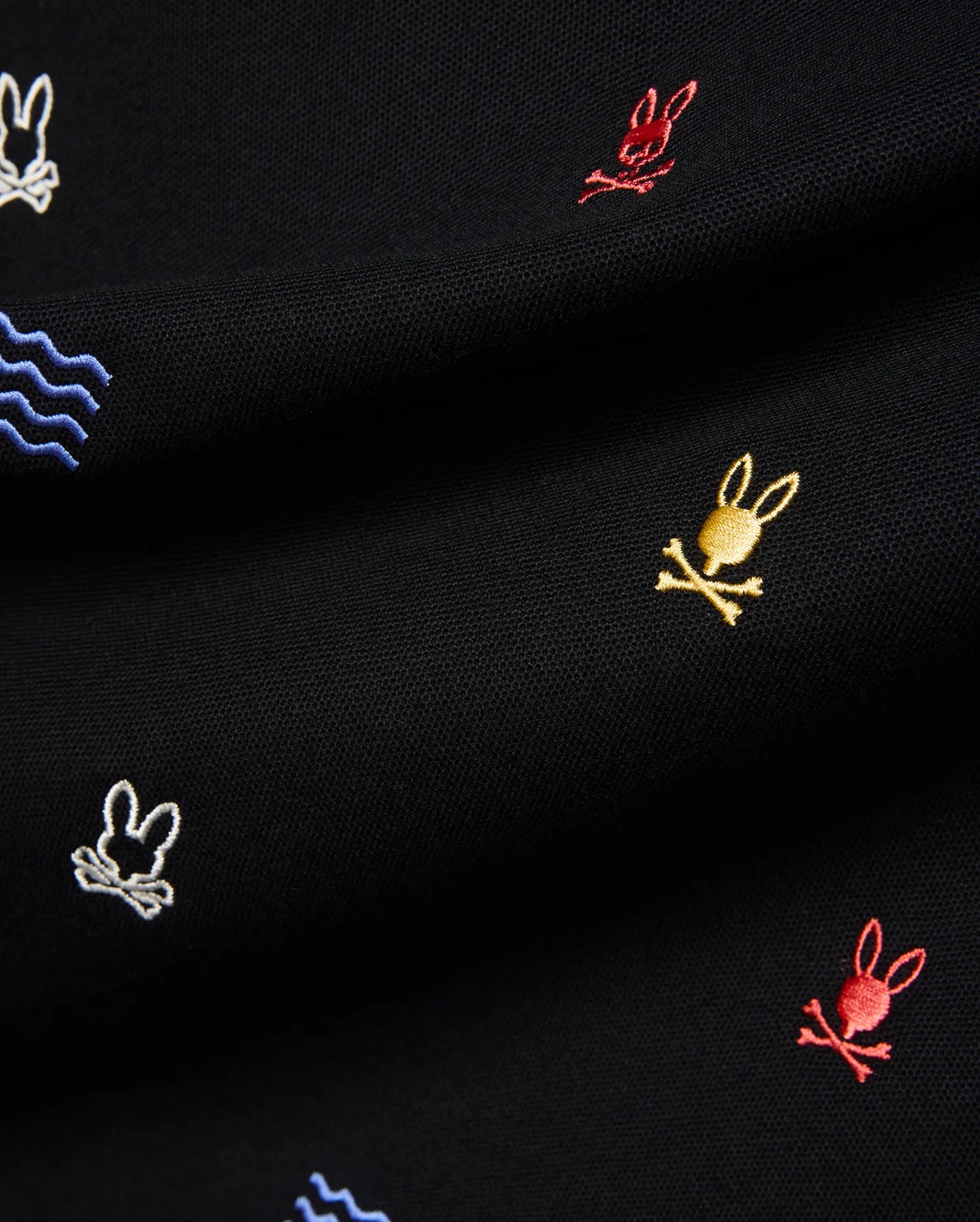Mens Colin All-Over Pique Polo in black, featuring a classic pique texture with an all-over embroidered pattern of small, colorful Psycho Bunny skull logos in red, yellow, and white, alongside blue wave motifs.