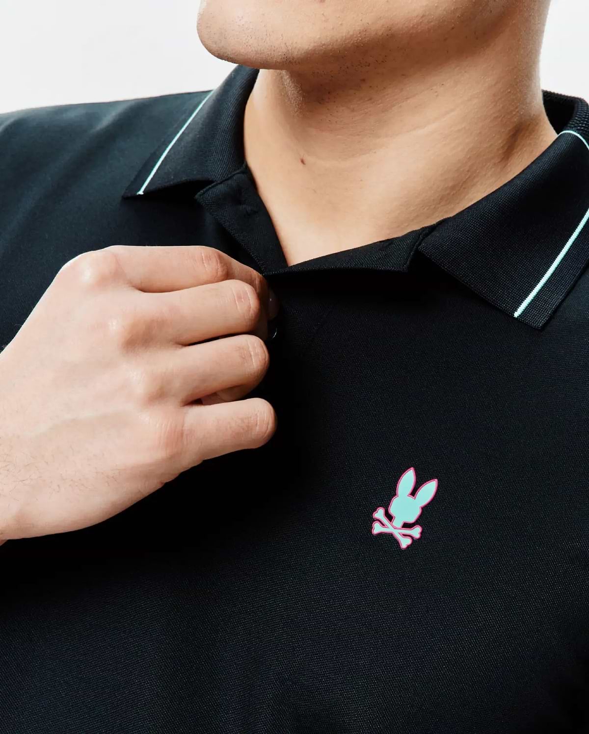Mens Edwin Sport Polo, black pique knit with a classic fit, featuring a light blue collar stripe and an embroidered mint green and pink Psycho Bunny skull logo on the left chest.