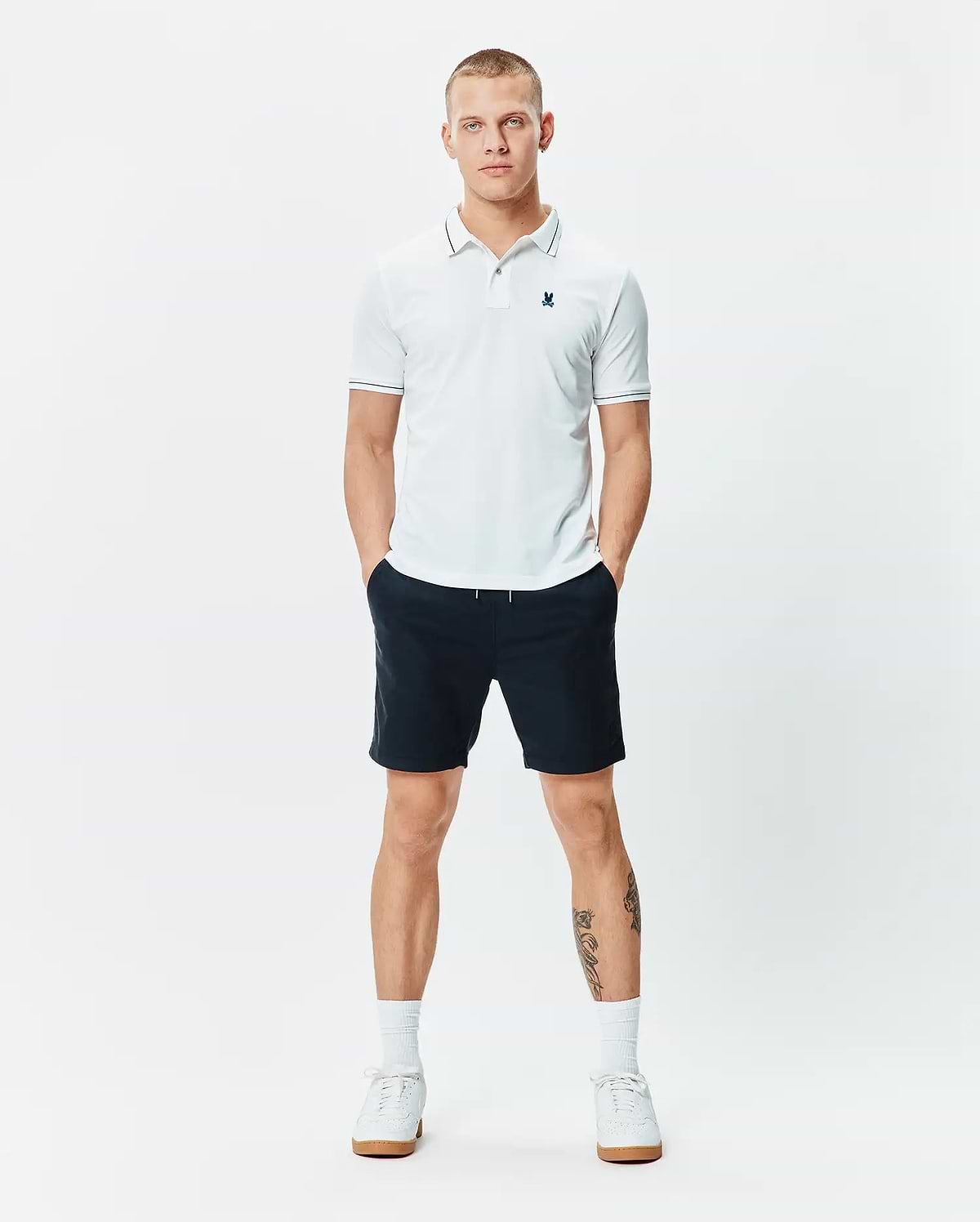 Mens Edwin Sport Polo, a classic fit white pique polo with navy tipping on the collar and cuffs, featuring a small embroidered teal Psycho Bunny skull logo on the left chest.